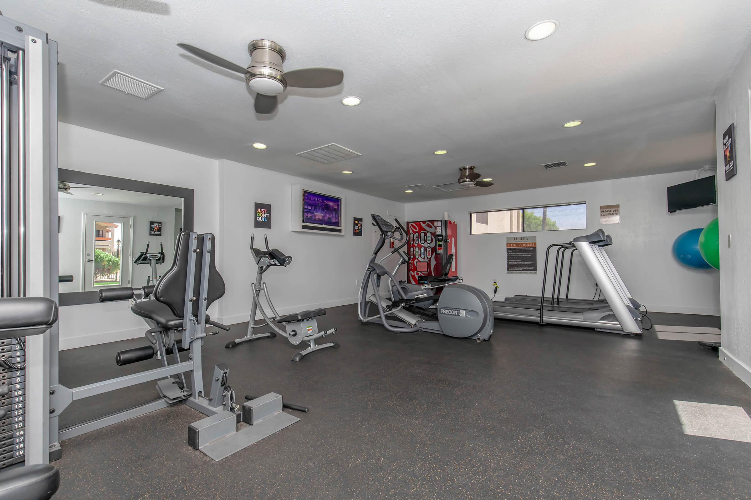 A modern gym interior featuring exercise equipment, including treadmills, elliptical machines, and weight machines. The space is well-lit with overhead lights and a ceiling fan, and includes a large mirror and fitness accessories like exercise balls, along with motivational posters on the walls.