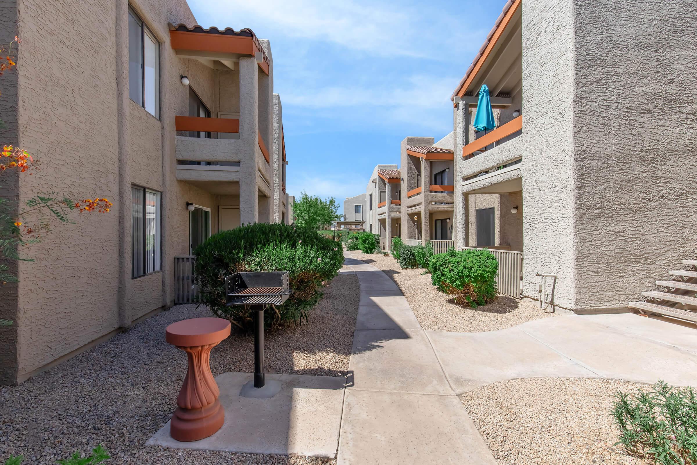 A view of an outdoor apartment complex featuring two-story buildings with balconies, surrounded by desert landscaping. A pathway runs through the middle, leading to a barbecue grill and lush greenery. Bright blue sky with scattered clouds overhead.