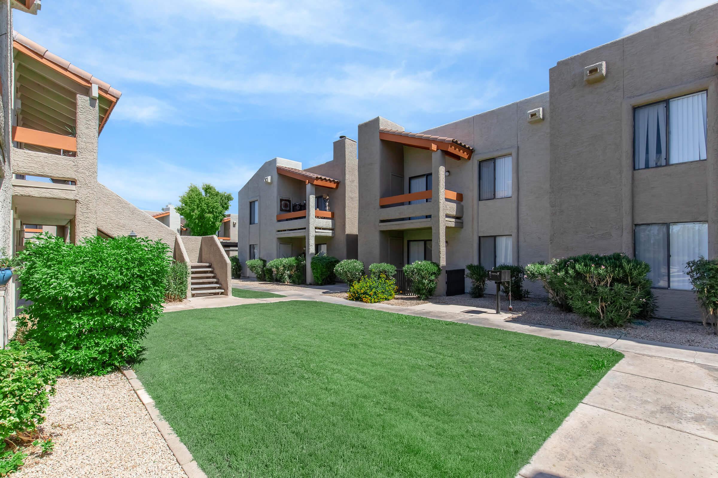 A landscaped courtyard view of an apartment complex featuring two-story buildings with balconies. There is lush green grass in the foreground, surrounded by shrubs and small trees. A set of stairs leads to the second floor, with clear blue skies above.