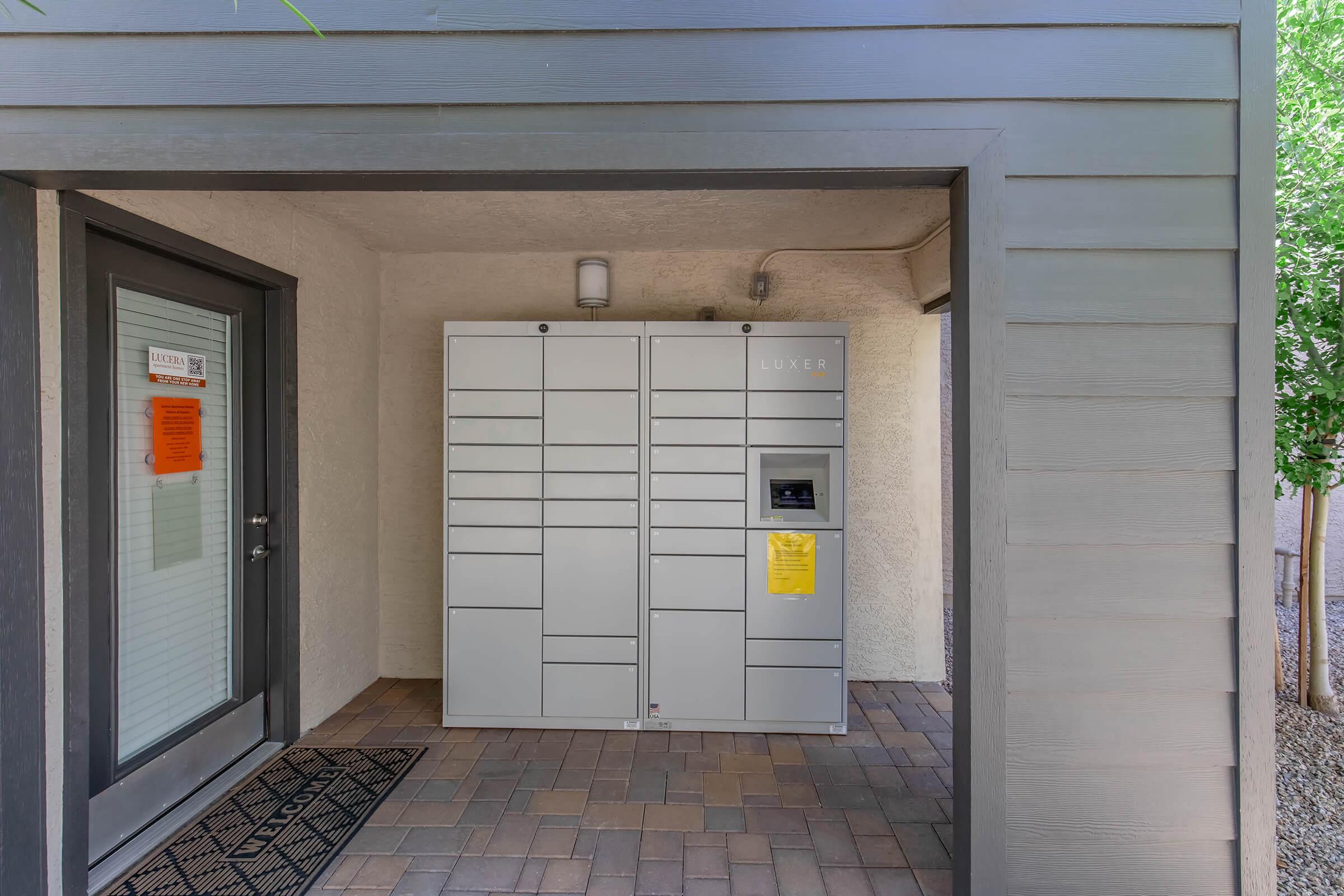 A modern package locker system situated in a covered entrance area. The lockers are gray and feature multiple compartments for secure package retrieval. There is a notice on the door and a mat at the entrance, with surrounding landscaping providing a neat and tidy appearance.