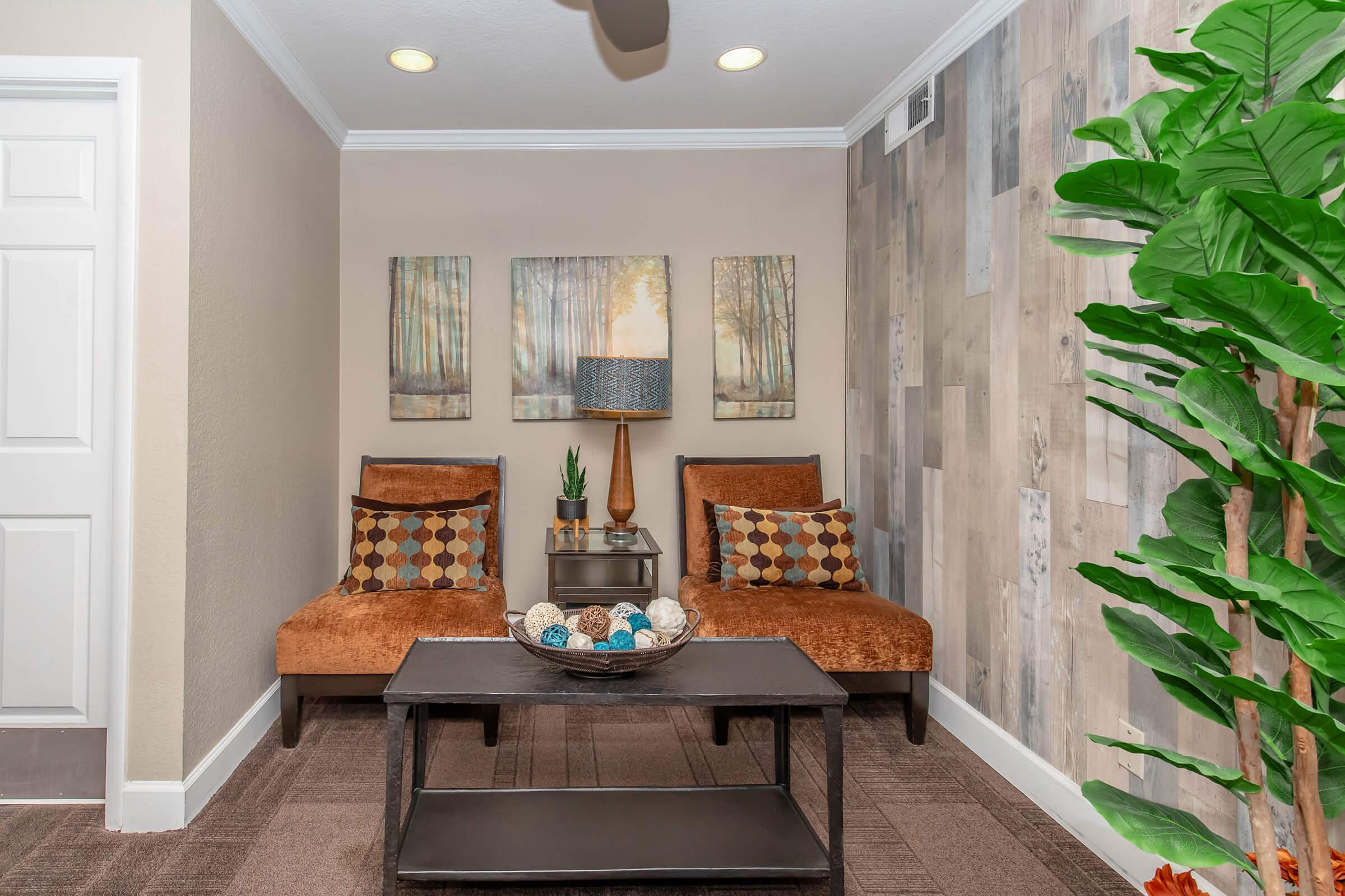 A cozy interior space featuring two brown armchairs with patterned cushions, a modern coffee table in the center, and a decorative bowl. The wall displays three nature-inspired artworks, and there's a stylish lamp on the table. A large green plant adds a touch of greenery to the room.