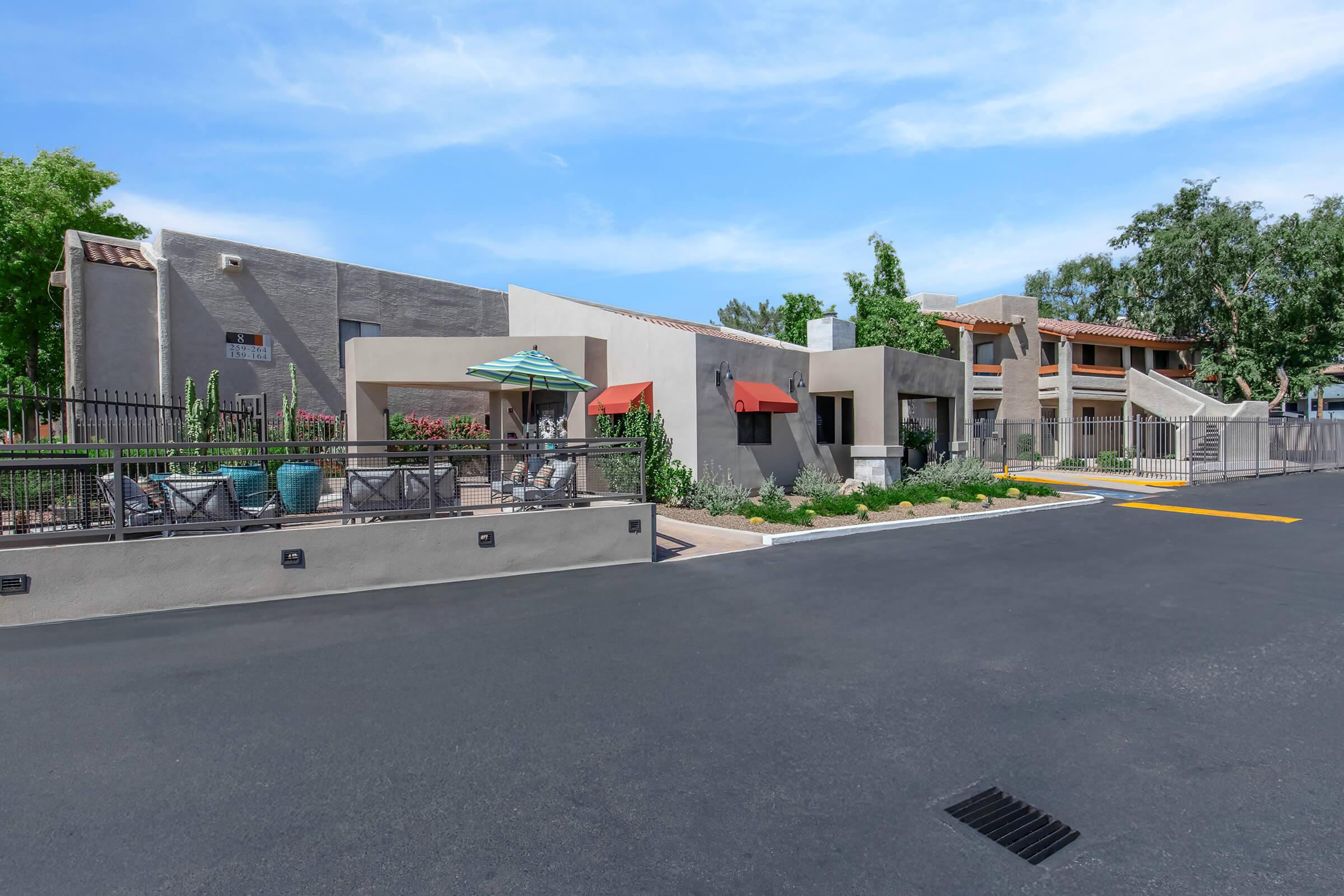 A view of a modern residential complex featuring landscaped gardens, outdoor seating areas with umbrellas, and two-story buildings. The entrance is gated, and the road is paved. The setting includes greenery and a clear blue sky, creating a welcoming atmosphere.