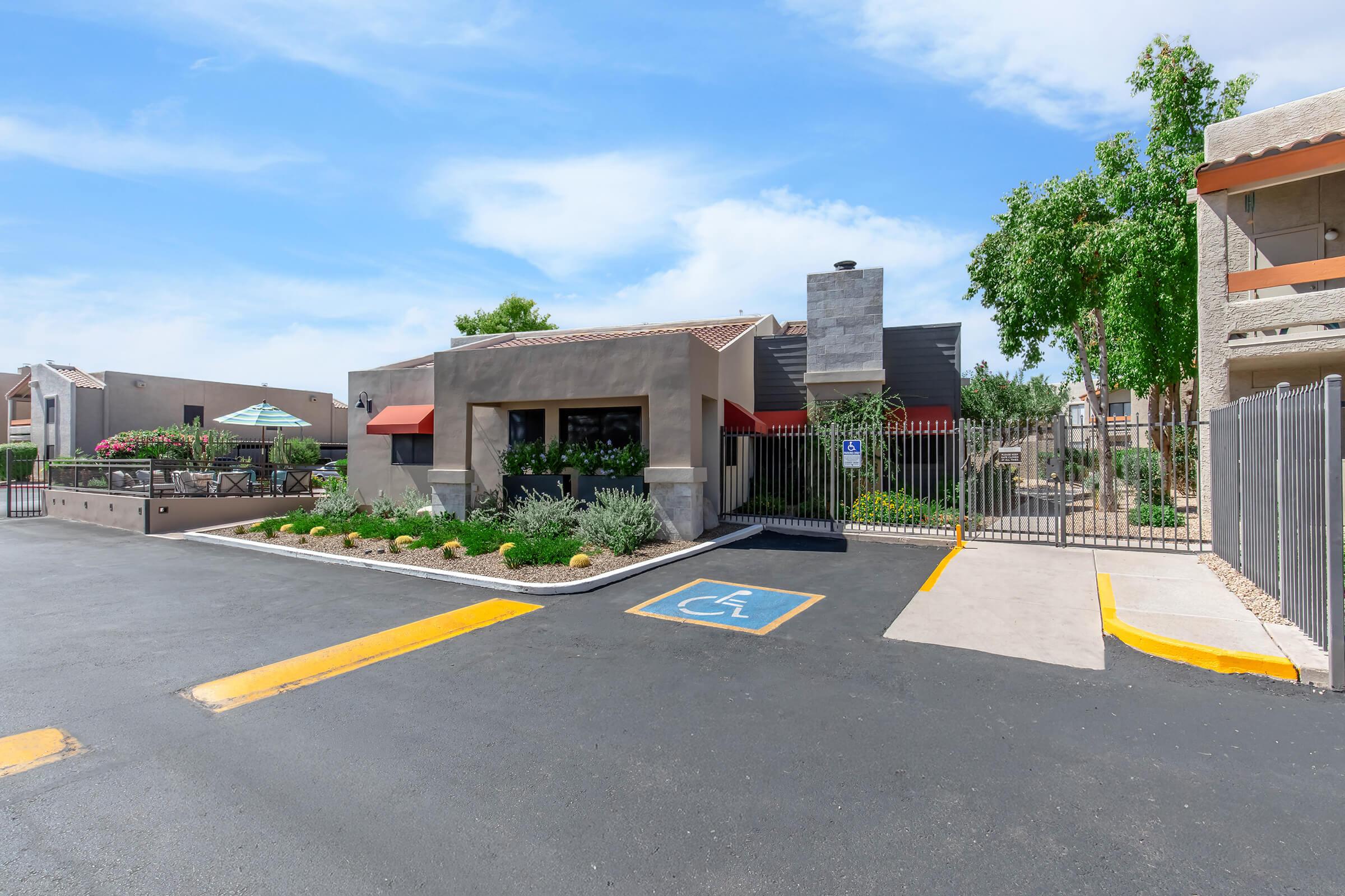 A well-maintained exterior of a residential complex featuring a reception area, landscaped greenery, and accessible parking. The building has a modern design with large windows, and the entrance is gated, enhancing security. Clear blue skies add brightness to the scene.