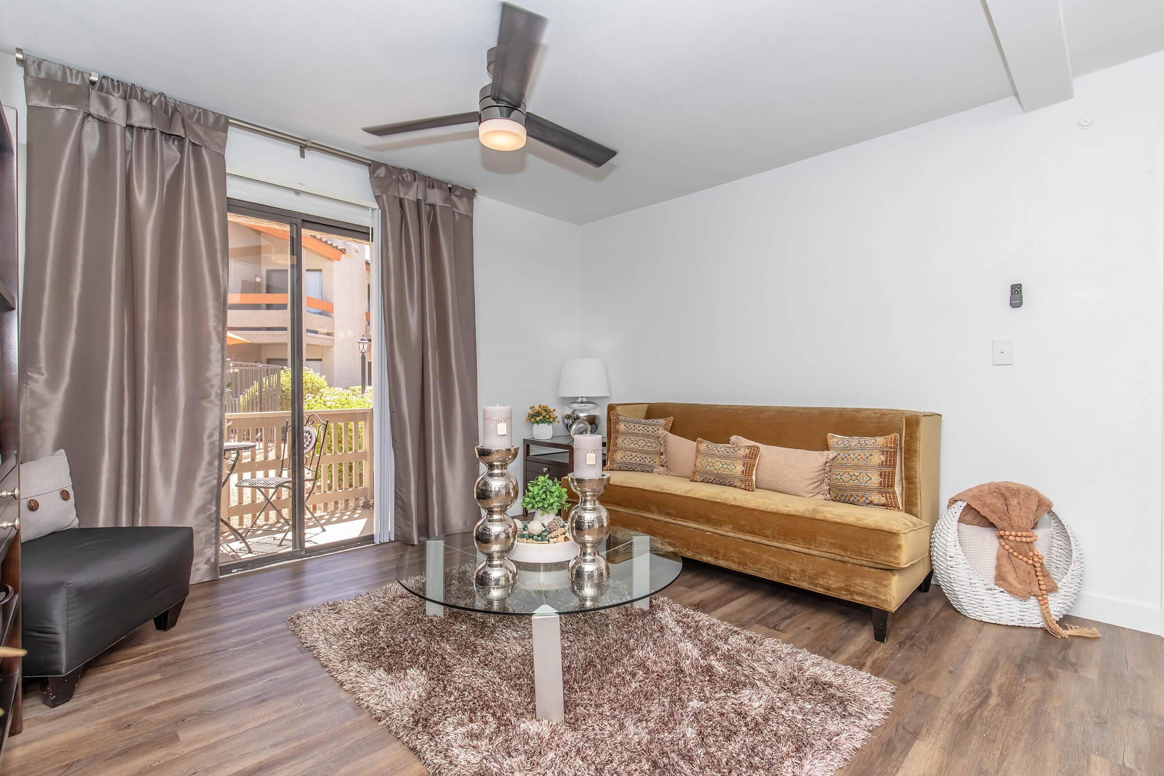 A modern living room featuring a cozy brown velvet sofa, a glass coffee table with decorative candles, and a plush area rug. The space is illuminated by natural light from a sliding glass door leading to a balcony, accented by elegant curtains and a stylish floor lamp.