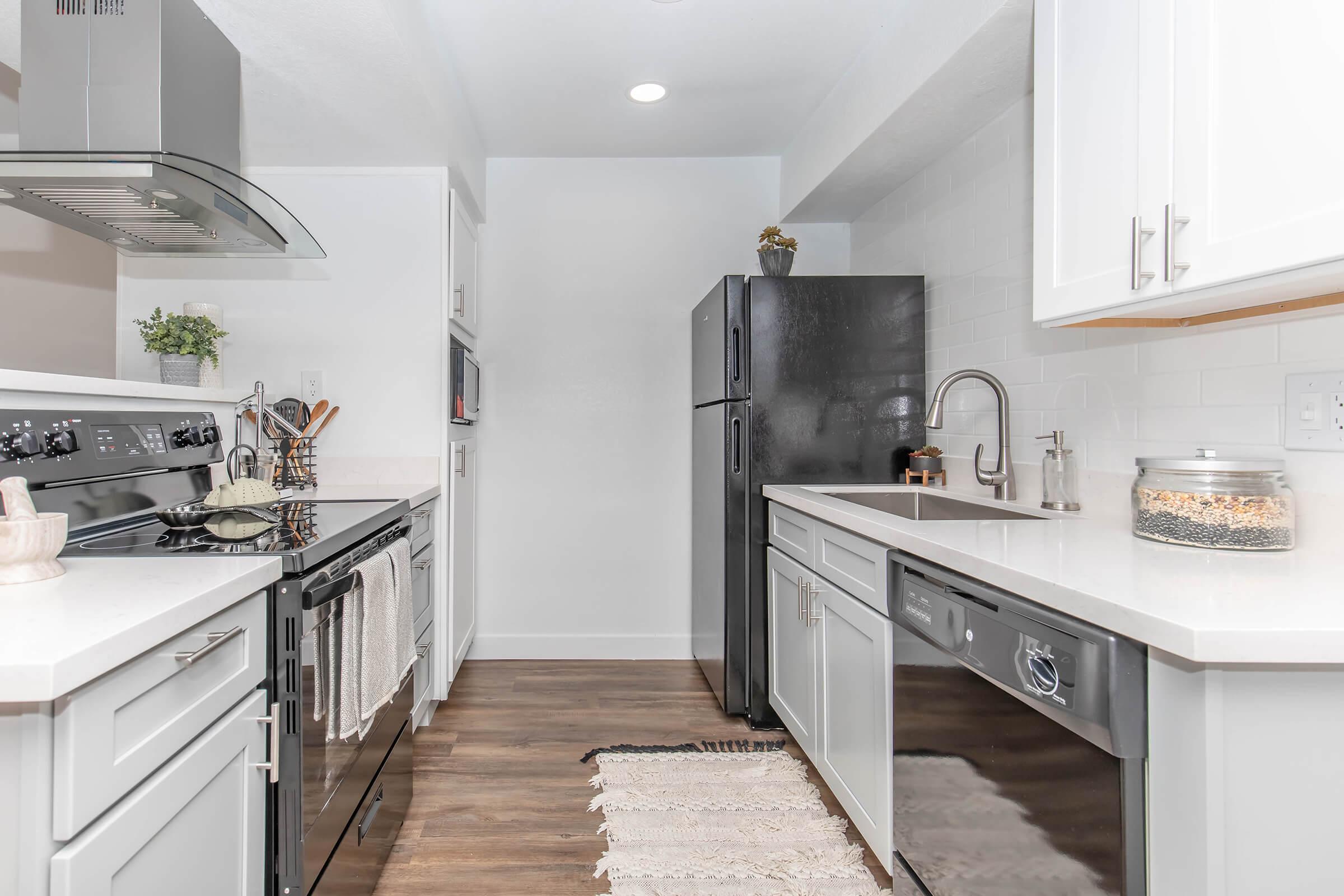 A modern kitchen featuring white cabinetry, stainless steel appliances, and a dark refrigerator. The countertops are a light color, with a rug on the floor and a small plant on the counter. Task lighting illuminates the space, creating a bright and inviting atmosphere.