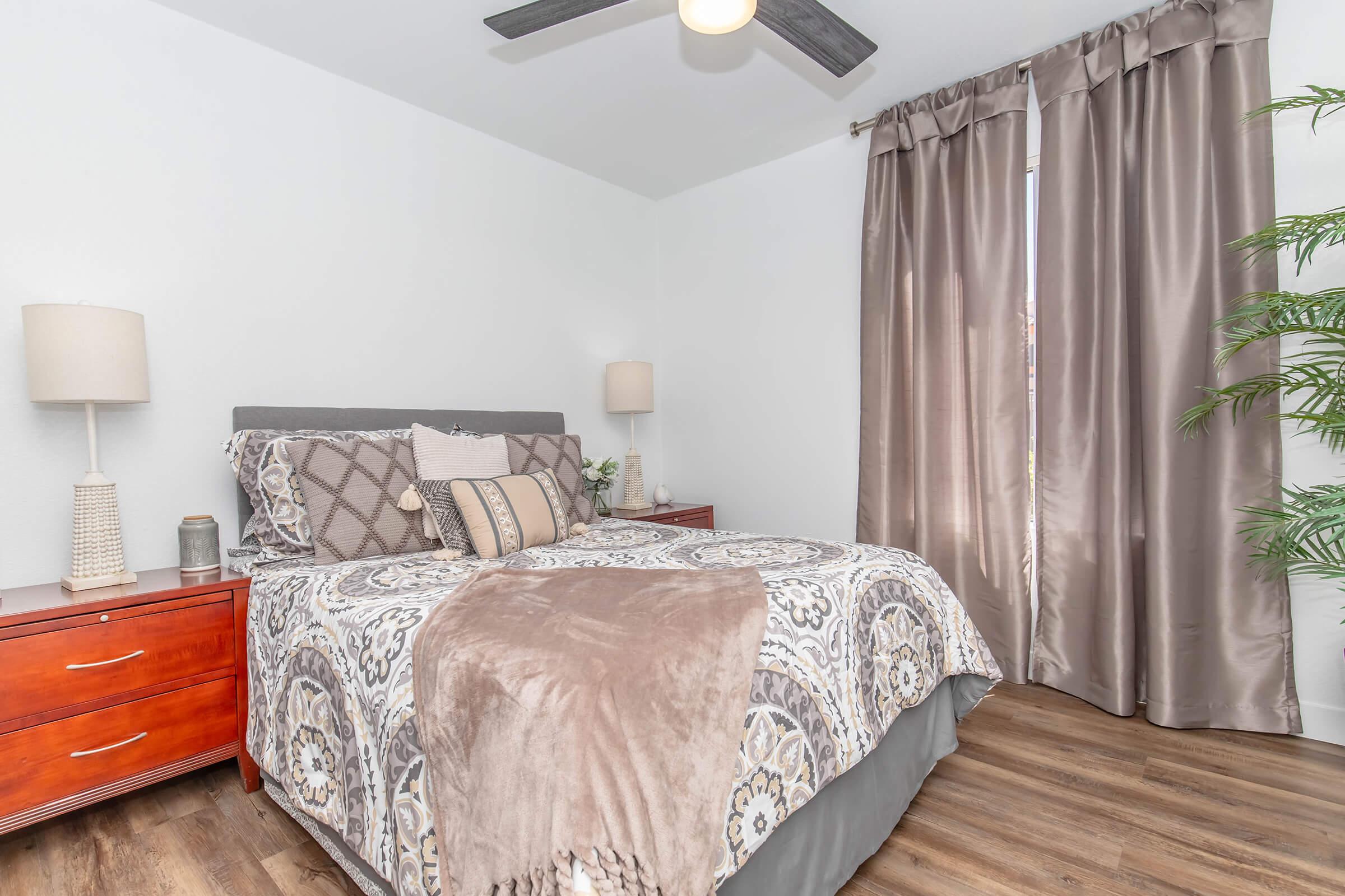 A cozy bedroom featuring a king-sized bed with patterned bedding and decorative pillows. Nightstands with lamps sit on either side of the bed. Dark curtains frame the window, and a potted plant adds a touch of greenery to the warm, inviting space with wooden flooring.