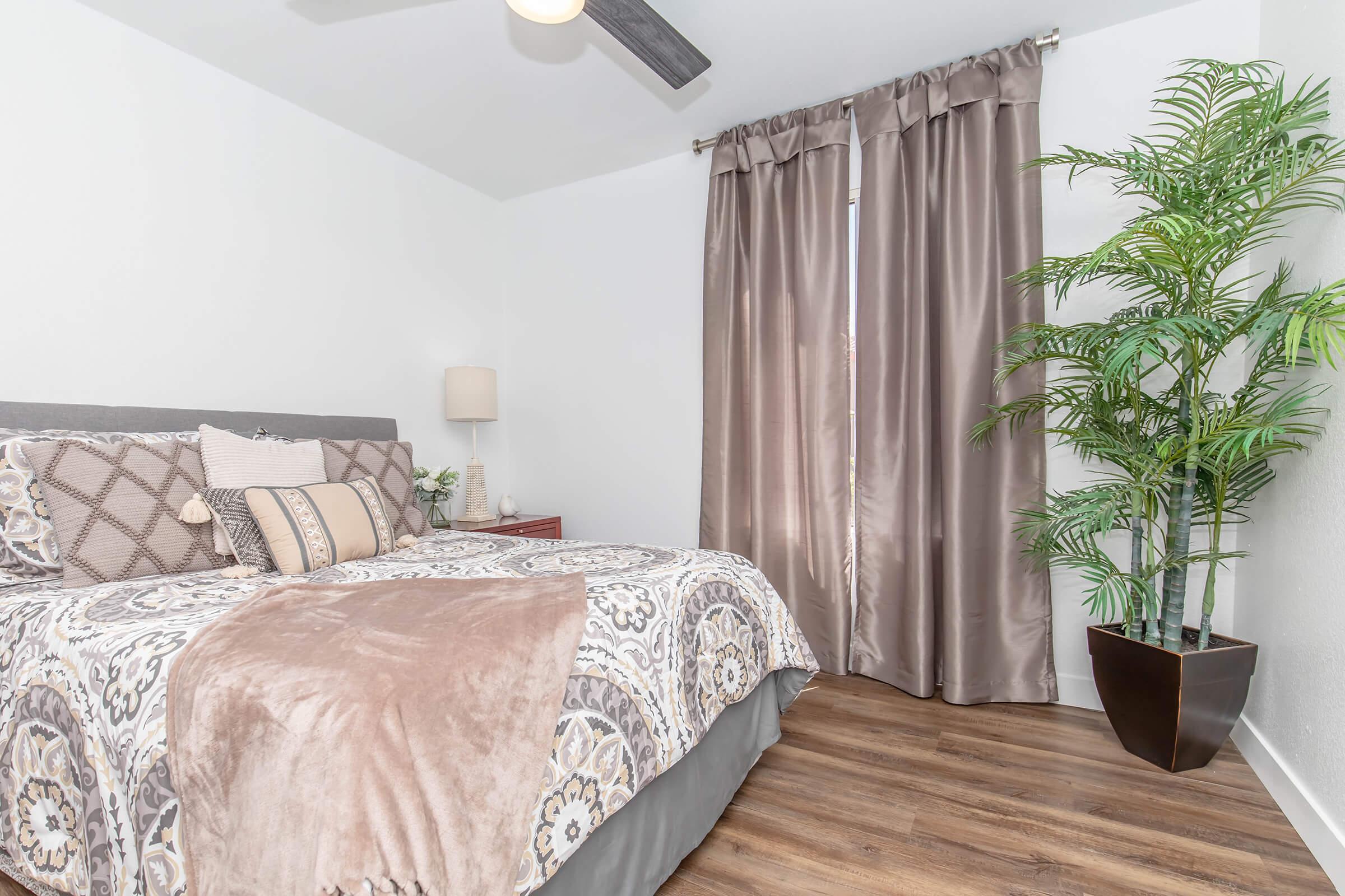 A cozy bedroom featuring a neatly made bed with decorative pillows and a patterned blanket. There are floor-to-ceiling curtains covering a window, a potted plant in the corner, and a bedside lamp on a small table, all set against a light-colored wall and wooden floor.