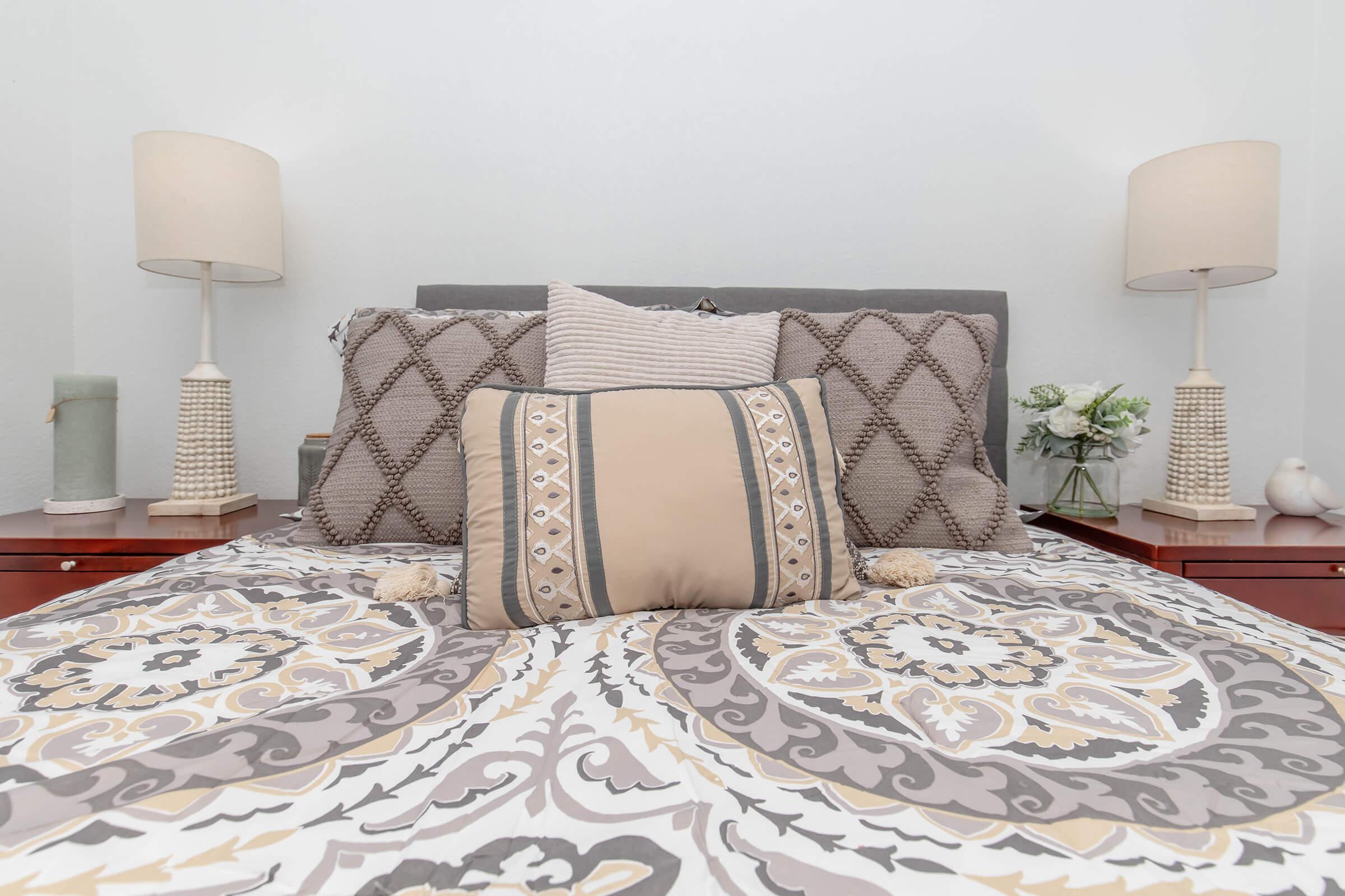 A neatly made bed with a decorative patterned comforter and multiple pillows arranged on a gray headboard. Two bedside lamps with textured bases are placed on wooden nightstands, which also feature small decorative items and a vase with flowers. The overall decor is warm and inviting.