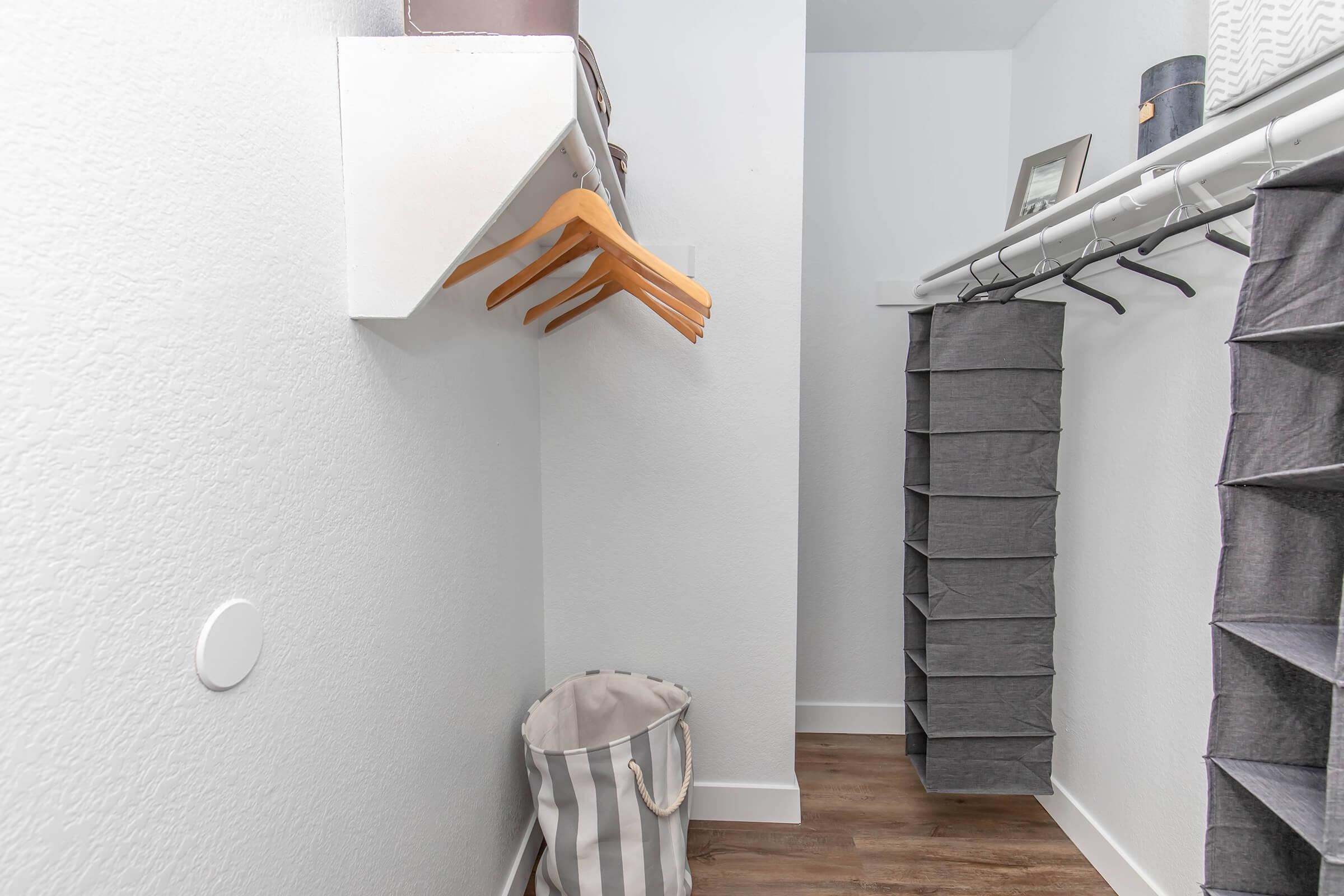 A neatly organized closet featuring a white wall with a shelf and wooden hangers on the left. The right side displays a hanging storage unit with gray fabric bins. A striped fabric bag sits on the floor, and the space has a clean, modern look with wooden flooring.