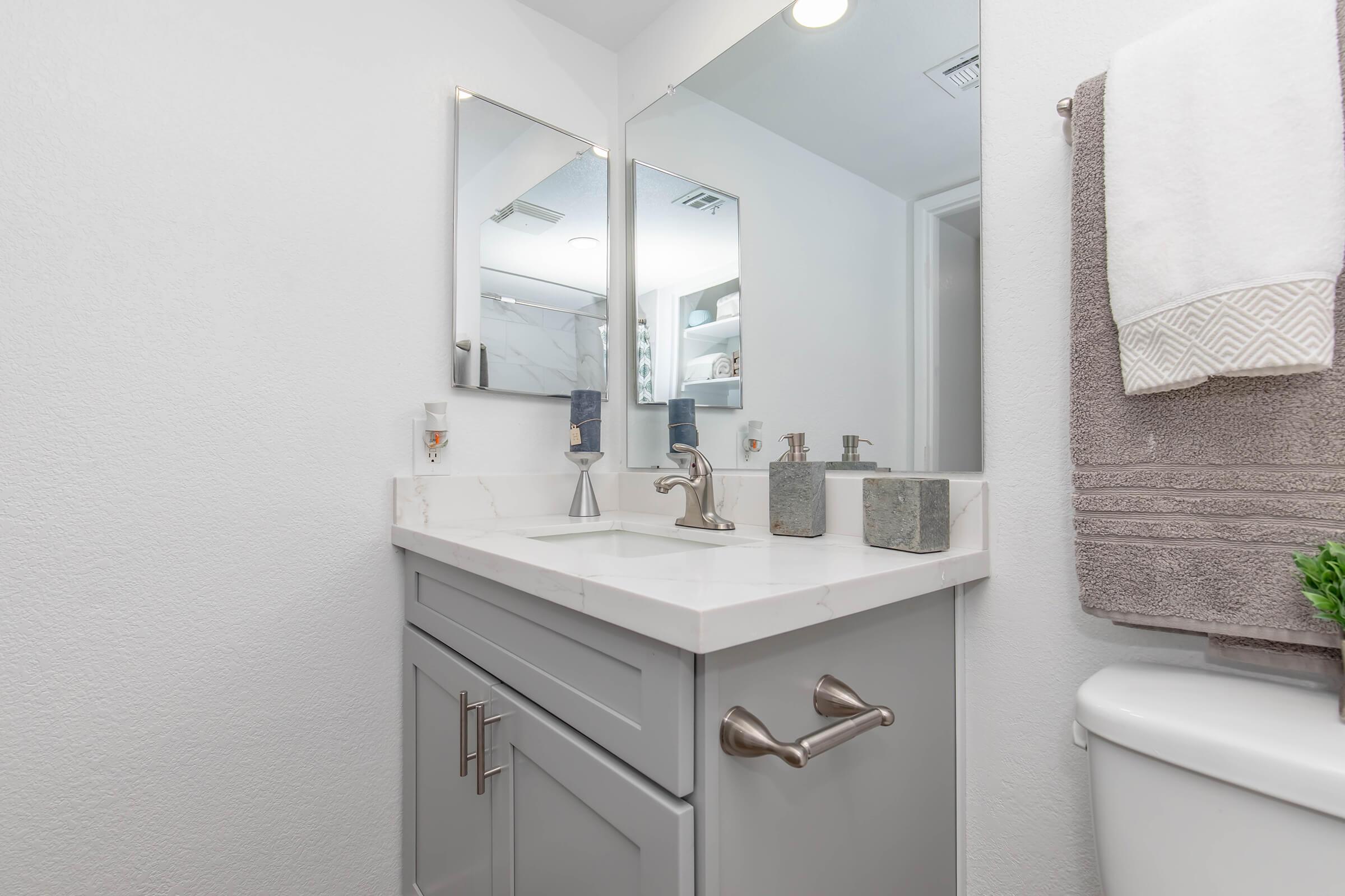 Modern bathroom with a light gray vanity featuring a white marble countertop, two mirrors above the sink, and stylish sink fixtures. Gray towels hang to the right, and a small plant adds a touch of greenery. The walls are painted white, creating a bright and clean atmosphere.