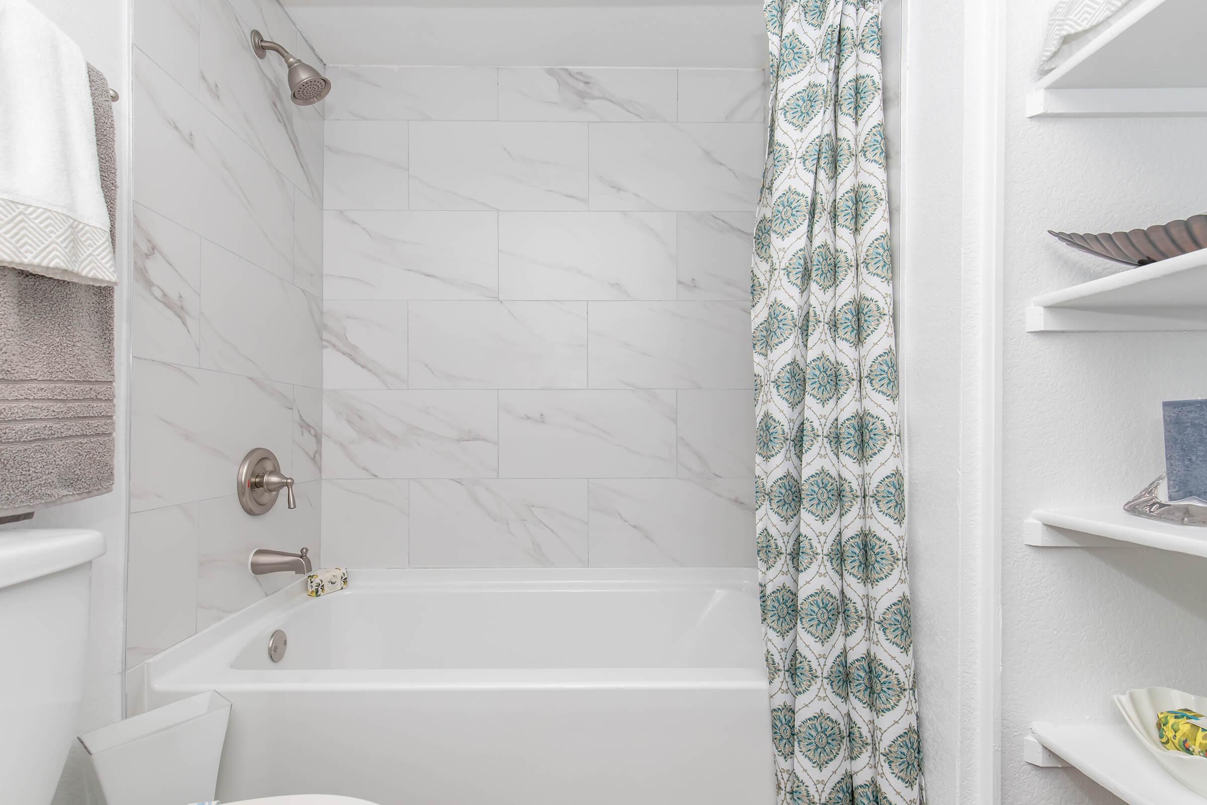 A modern bathroom featuring a bathtub with a showerhead, white tiled walls, and a patterned shower curtain. There are towels hanging nearby and a shelf with decorative items, creating a clean and minimalistic aesthetic.