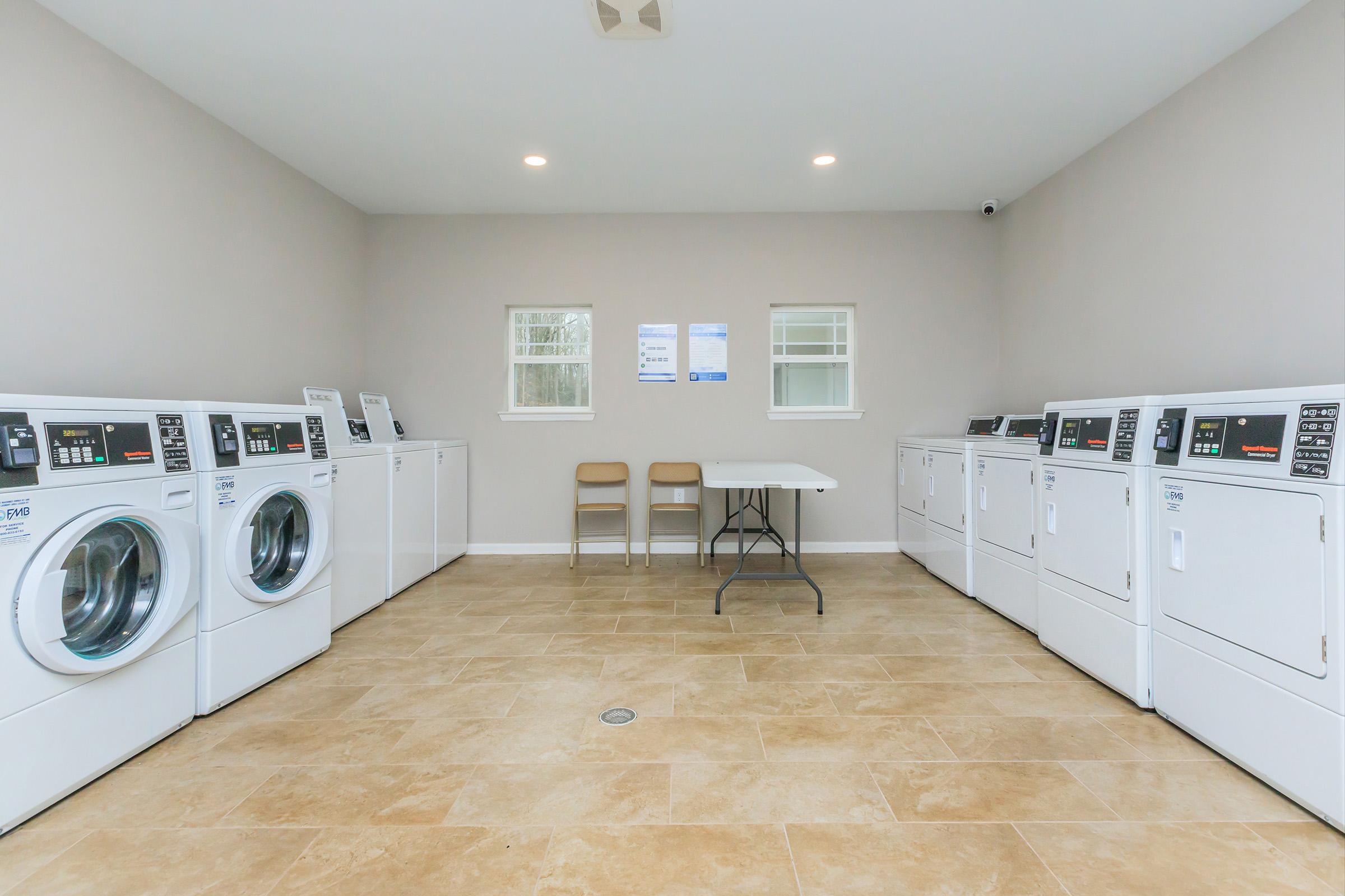 A clean, well-lit laundry room featuring multiple washing machines and dryers lined up against the walls. In the center, there is a simple table with two chairs. The walls are painted in a light color, and there are windows allowing natural light to enter the space.