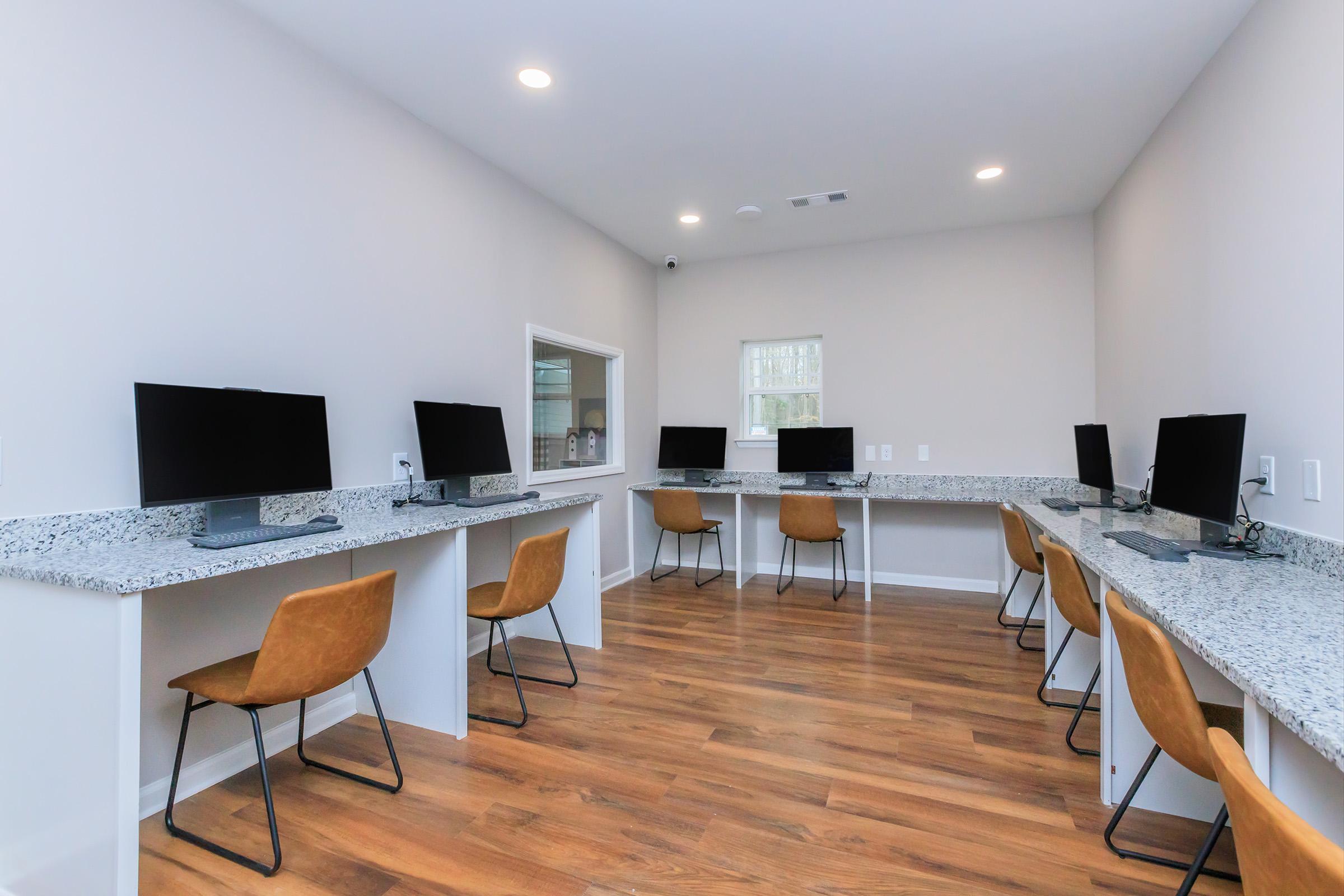 A modern office space featuring multiple computer stations with black monitors on granite countertops. Each station has a brown chair, and the room has hardwood flooring and neutral-colored walls, with natural light coming through a window.