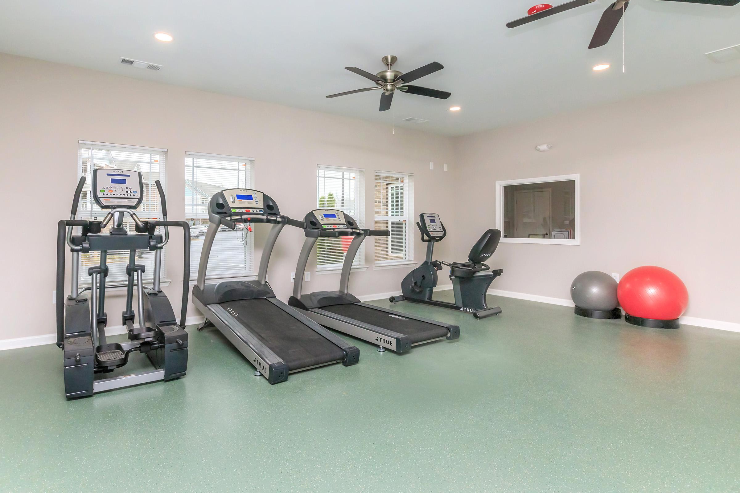 A modern gym with three treadmills and an elliptical machine, a stationary bike, and exercise balls. The room features large windows allowing natural light, ceiling fans, and green flooring, creating a bright and inviting workout space.