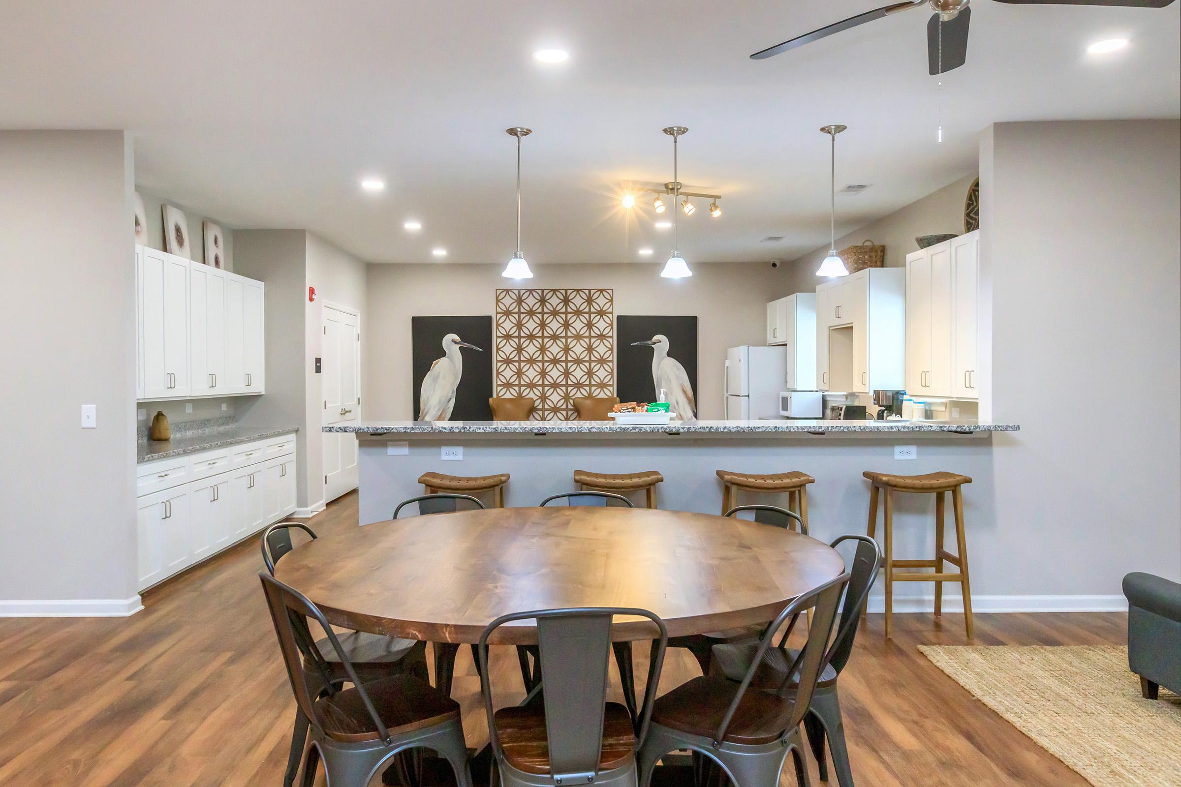 A modern kitchen and dining area featuring a large round wooden table surrounded by metal chairs, white cabinetry, and a granite countertop. The space is bright with a tray ceiling and elegant lighting fixtures, and two decorative heron sculptures on the wall add a stylish touch.