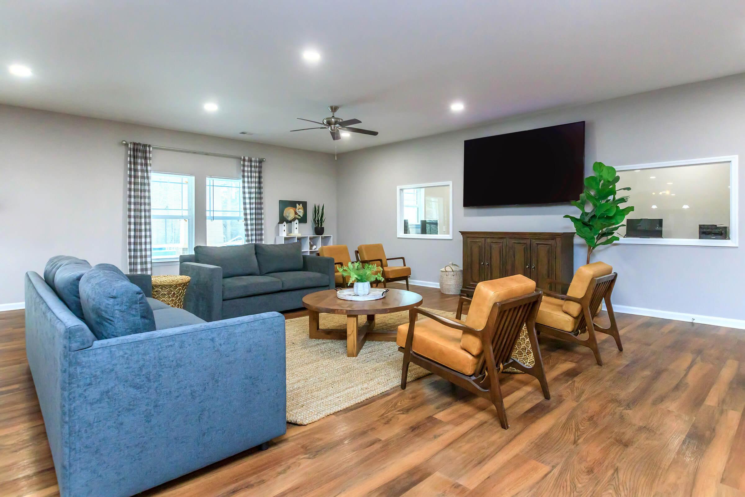A modern living room featuring a blue sofa, gray loveseat, and two brown armchairs arranged around a round coffee table. There’s a large flat-screen TV mounted on the wall and a potted plant in the corner. Natural light comes through windows adorned with checkered curtains, highlighting the warm wooden flooring.