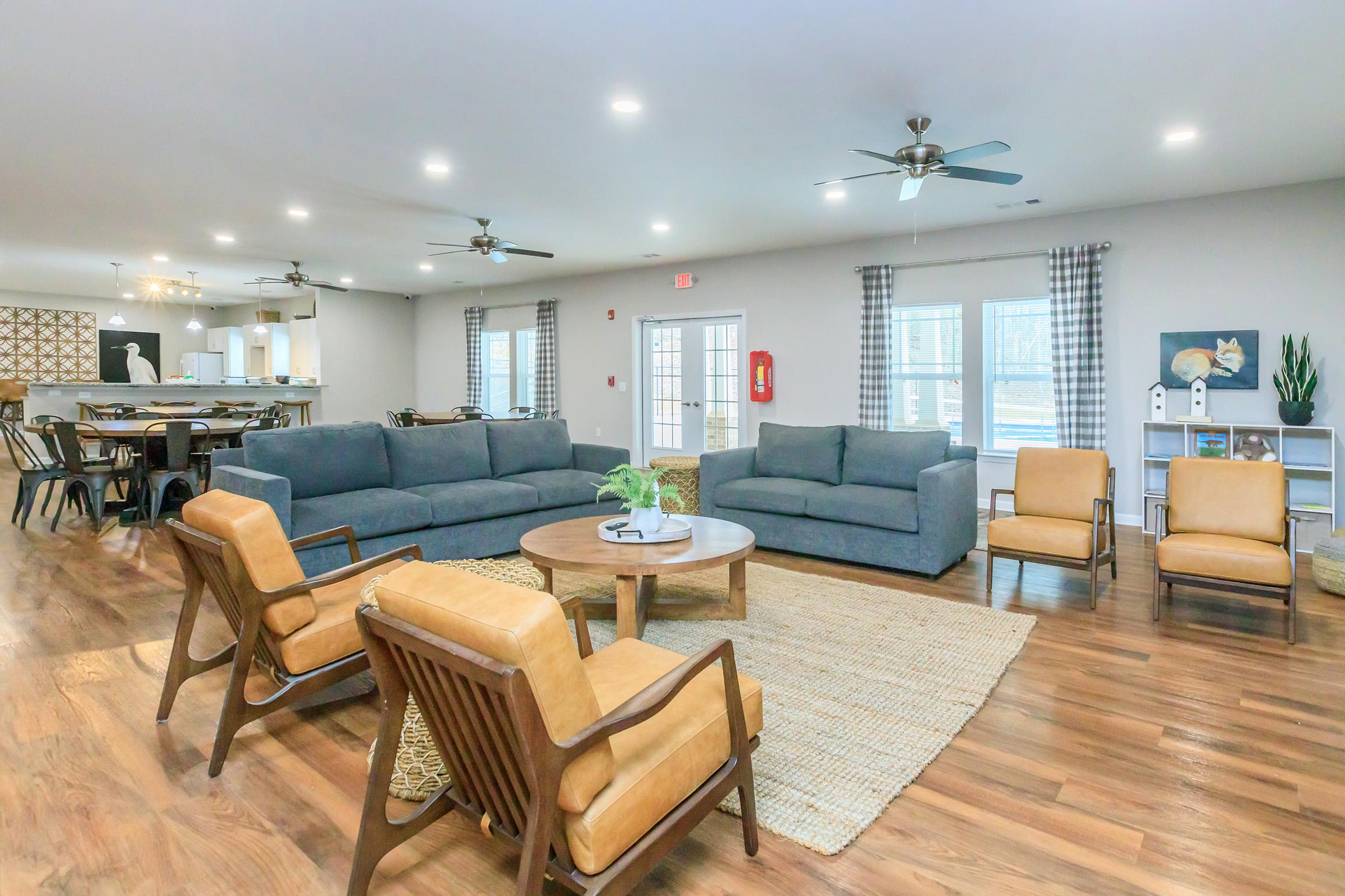 A spacious interior lounge area featuring two large gray sofas, comfortable tan armchairs, a round wooden coffee table, and a natural fiber rug. The room has light wood flooring, ceiling fans, and large windows with soft curtains, creating a cozy and inviting atmosphere. A dining area can be seen in the background.