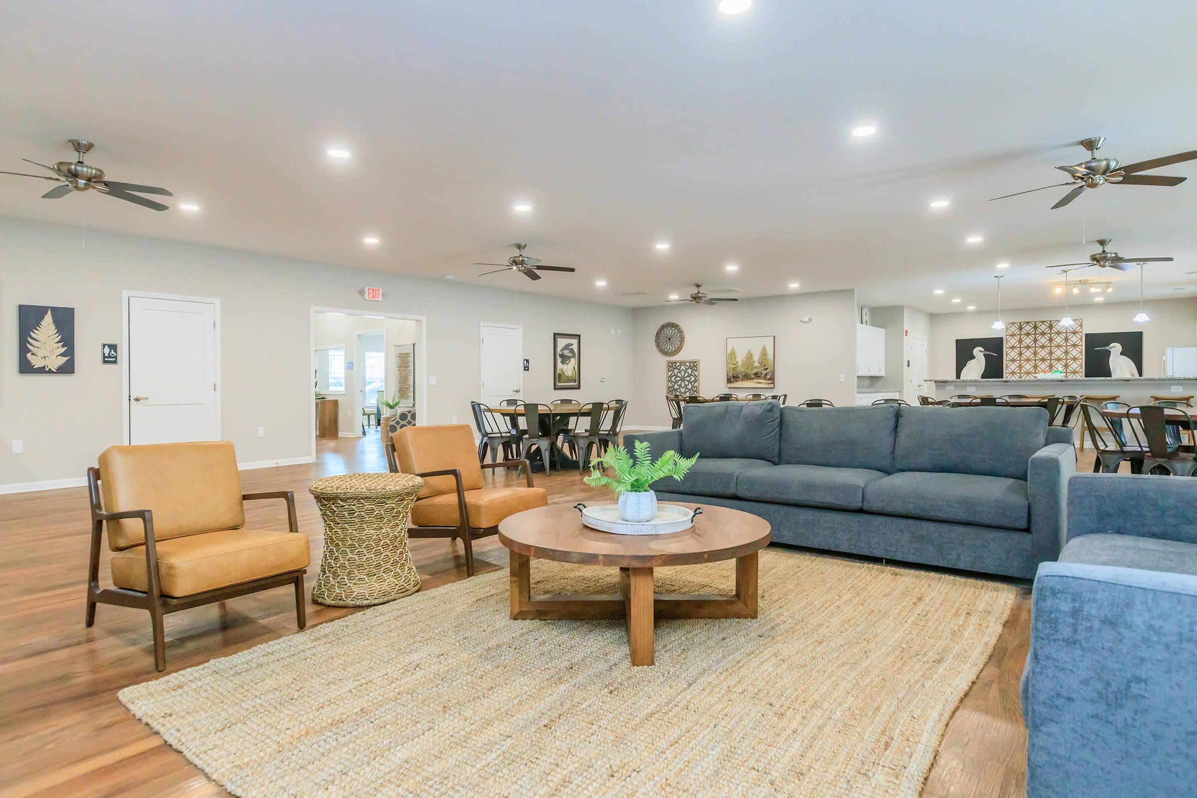 A spacious living area featuring a gray sofa, two brown chairs, and a round wooden coffee table on a large woven rug. The room is well-lit with ceiling fans and has decorative wall art. In the background, there is a dining area with tables and chairs, and the kitchen is visible with modern appliances.