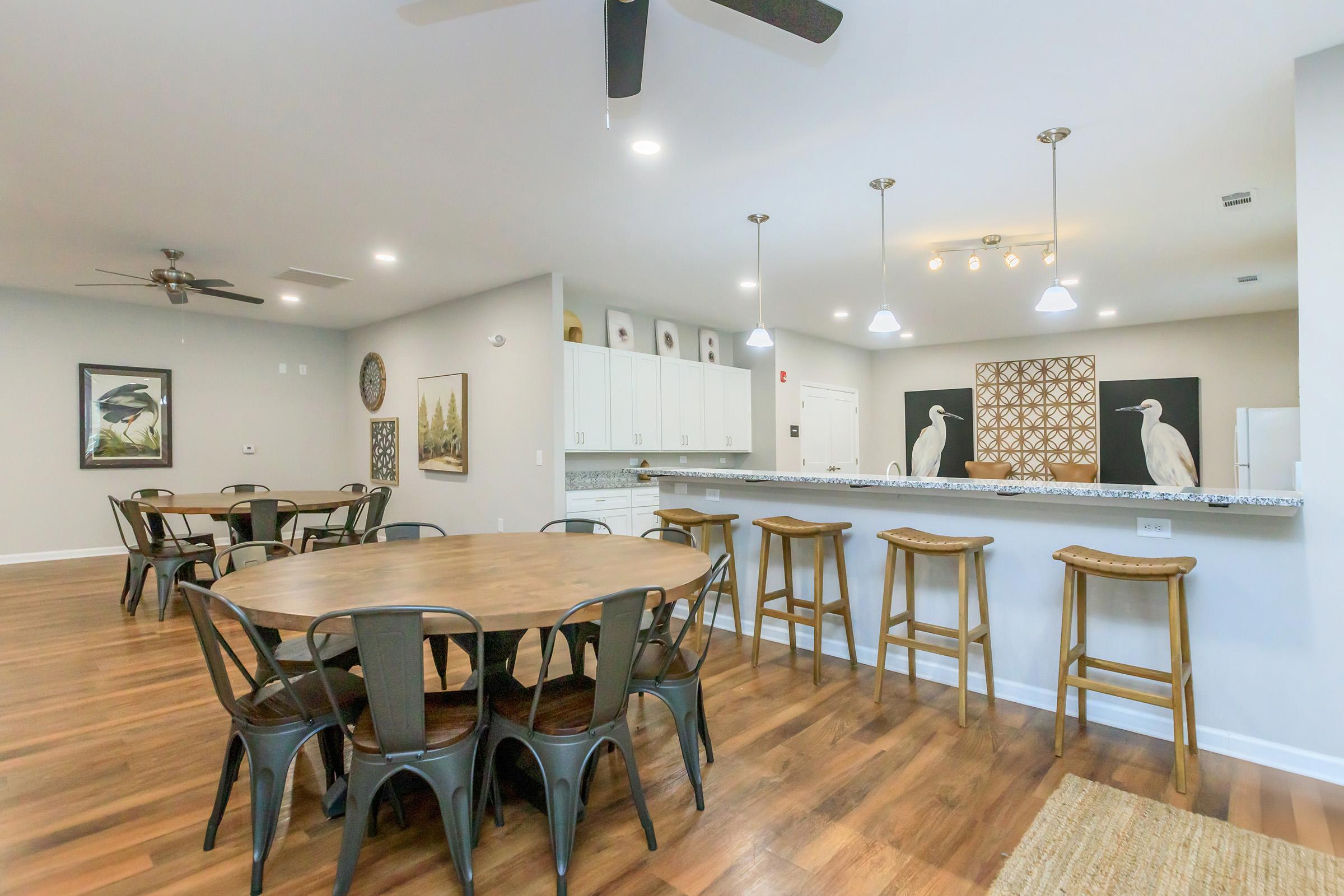 A spacious, modern dining area featuring a large round wooden table surrounded by metal chairs. A bar with stools is visible in the background, along with white cabinetry and decorative wall art. The room is well-lit with ceiling fans and bright lighting, creating a welcoming atmosphere.