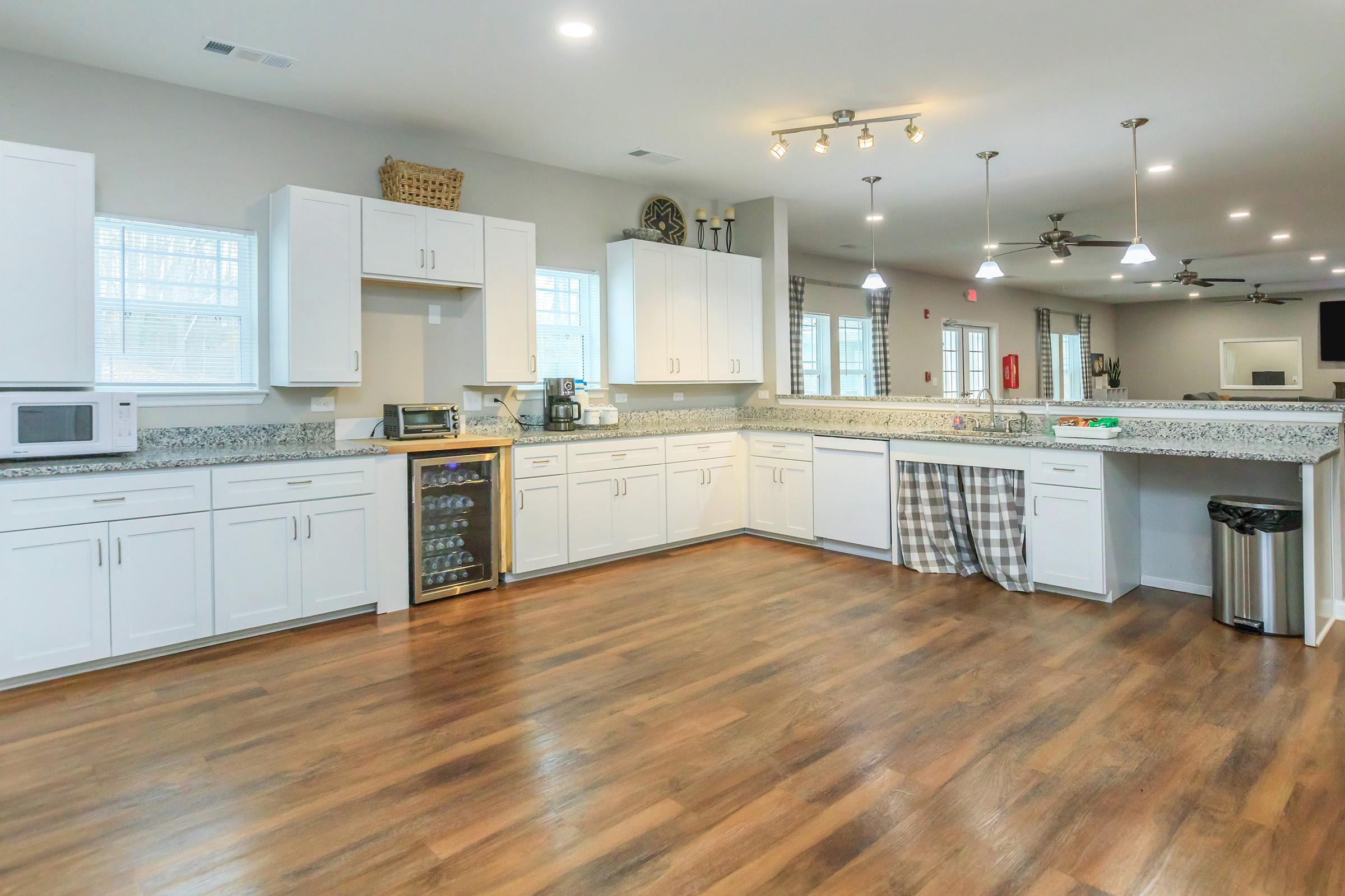 A modern kitchen featuring white cabinets, granite countertops, and wood flooring. It includes a microwave, small refrigerator, and a wine cooler. The space is well-lit with ceiling lights and has large windows, offering an open and inviting atmosphere. A checkered curtain adds a decorative touch.