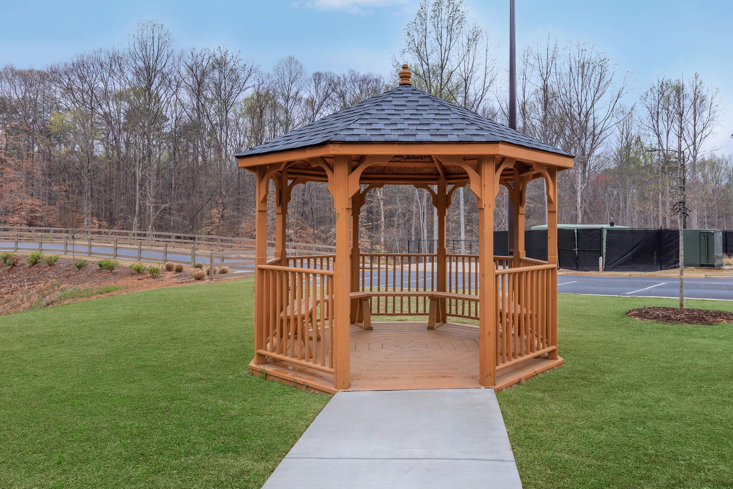 A wooden gazebo with a shingled roof, situated on a well-manicured lawn. Surrounding the gazebo are bare trees in the background and a paved walkway leading up to it. The scene is peaceful and inviting, suggesting a place for relaxation or gatherings.