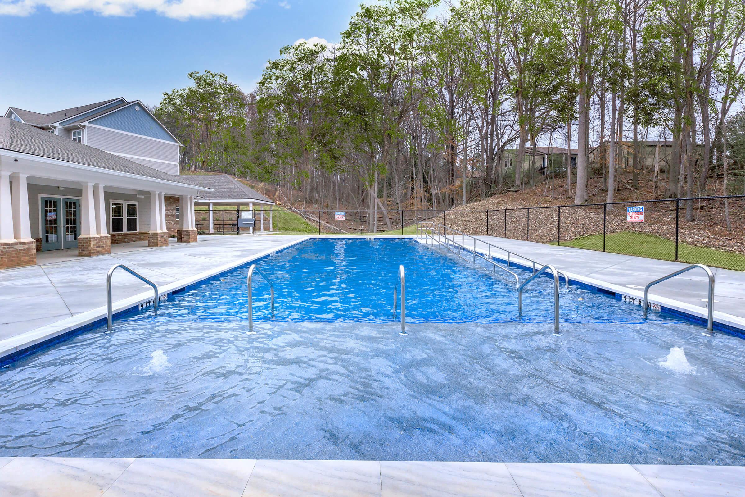A clear blue swimming pool surrounded by a patio area, with steps and railings leading into the water. Lush green trees are visible in the background, and a fenced area encloses the pool space. The sky is partially cloudy, creating a serene outdoor setting.