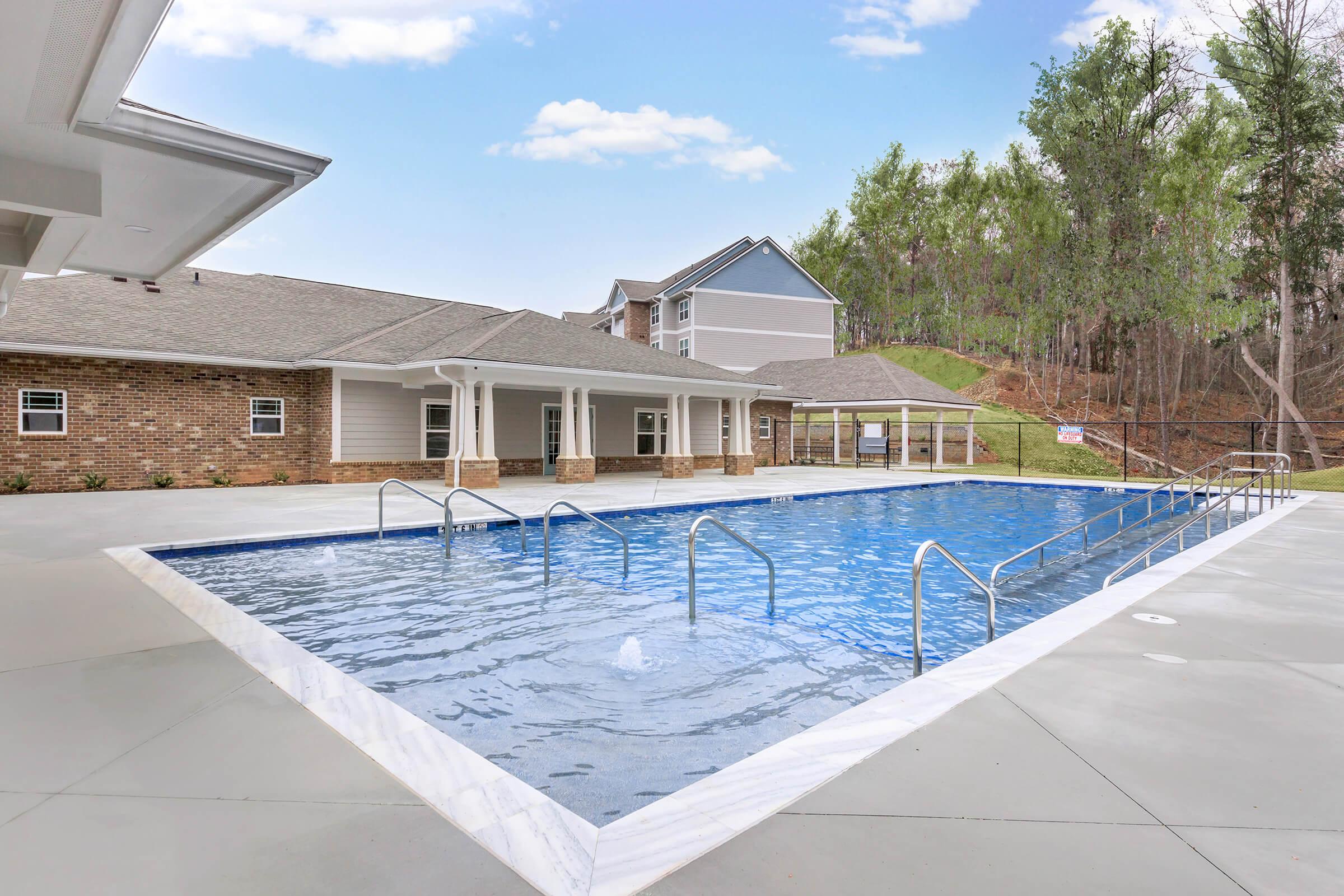 A clear blue swimming pool surrounded by a patio, featuring safety rails and a serene backdrop of trees. In the background, there are modern residential buildings with a light-colored exterior. The area is well-lit and inviting, ideal for relaxation and leisure activities.