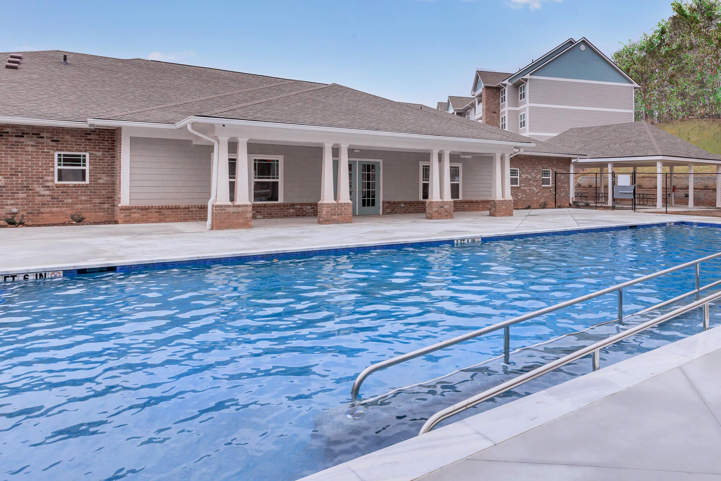 A clear blue swimming pool with a spacious surrounding area, featuring a brick building with large columns in the background. The setting includes lounge areas and greenery in the distance, offering a relaxing atmosphere. The sky is bright and blue, enhancing the inviting feel of the outdoor space.