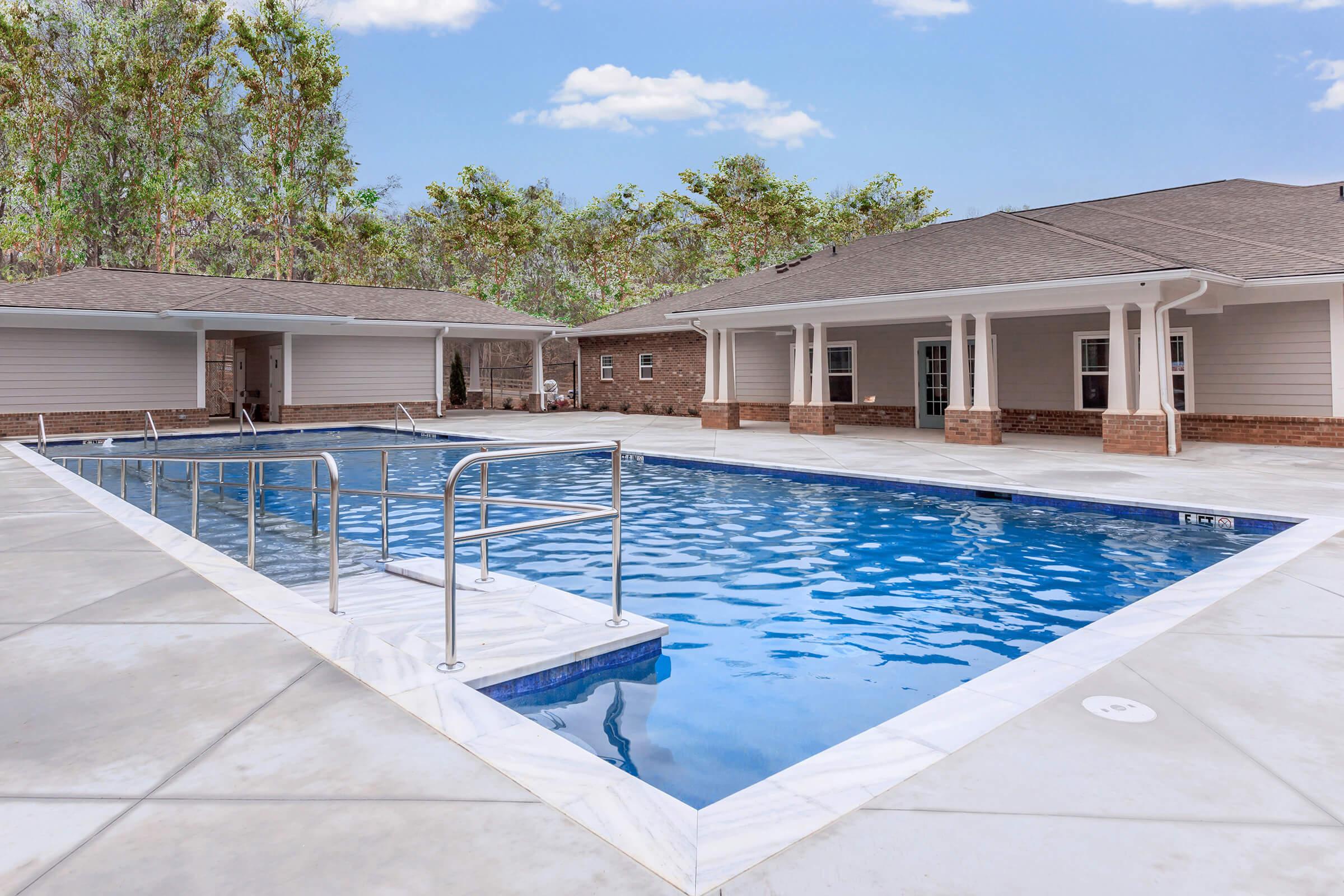 A spacious outdoor pool surrounded by a concrete deck, with a set of stairs leading into the water. Two residential buildings with light-colored siding and dark roofs are visible in the background, framed by trees. The sky is clear with a few clouds, creating a serene atmosphere.