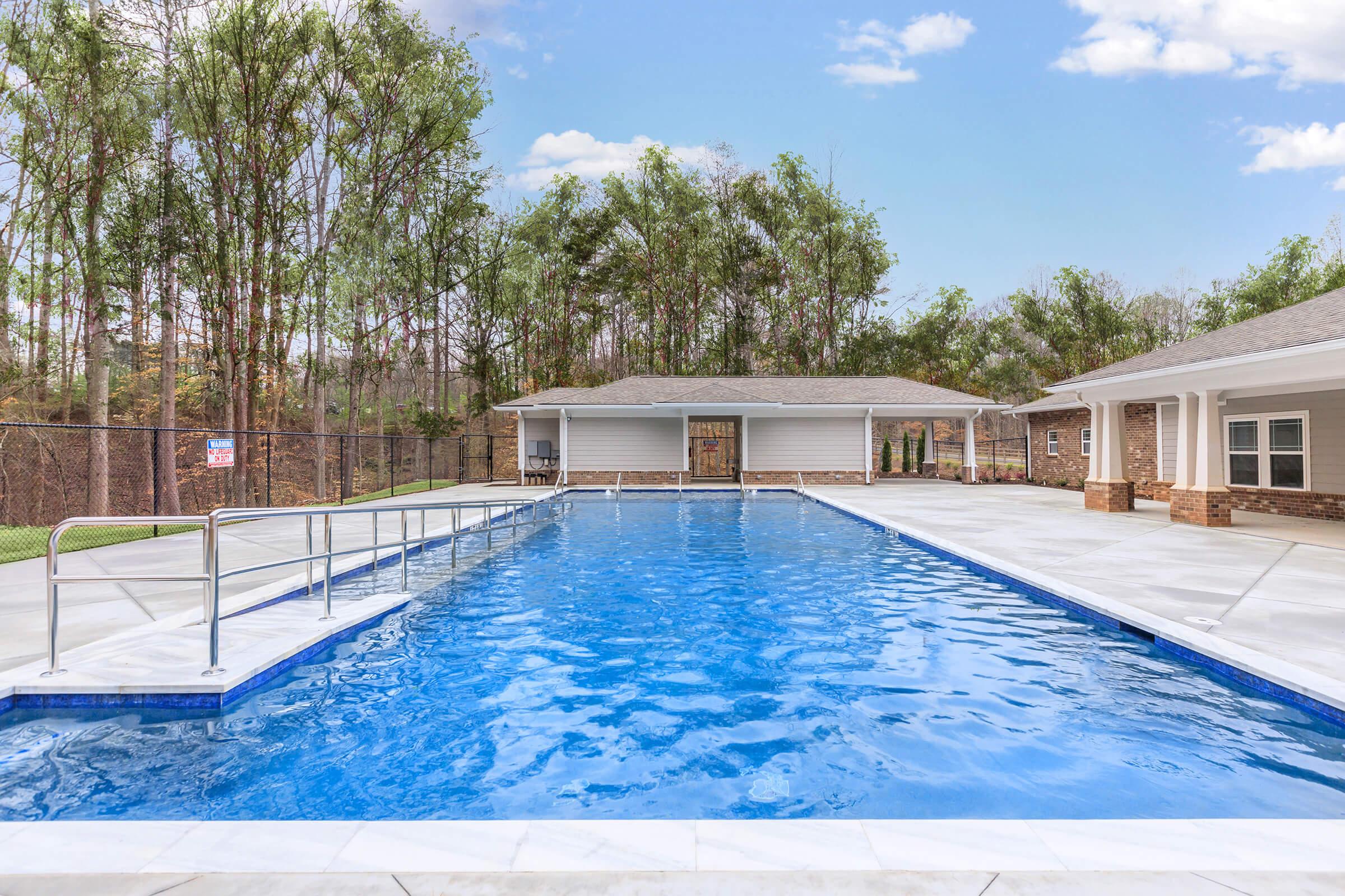 A clear blue swimming pool surrounded by a spacious deck. The pool features handrails for accessibility, with a wooded area visible in the background. Two buildings are situated nearby, one of which has large windows and a covered entrance. A chain-link fence encloses the area.
