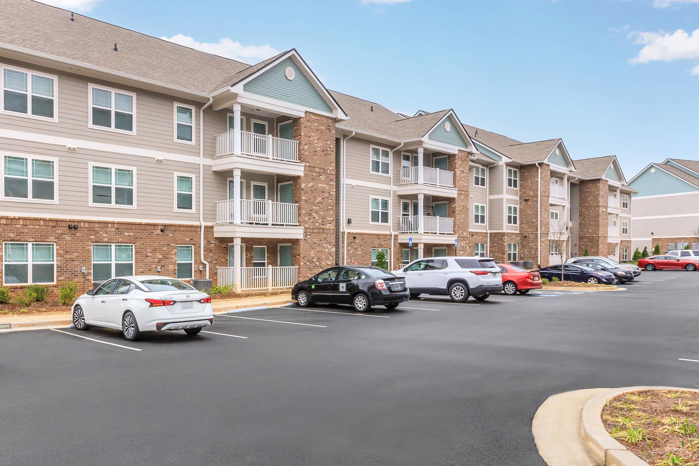 A multi-story apartment complex featuring light-colored siding and brick accents. Several parked cars are visible in the foreground, along with landscaped areas. The building has balconies and a well-maintained asphalt parking lot. A blue sky with a few clouds is visible in the background.