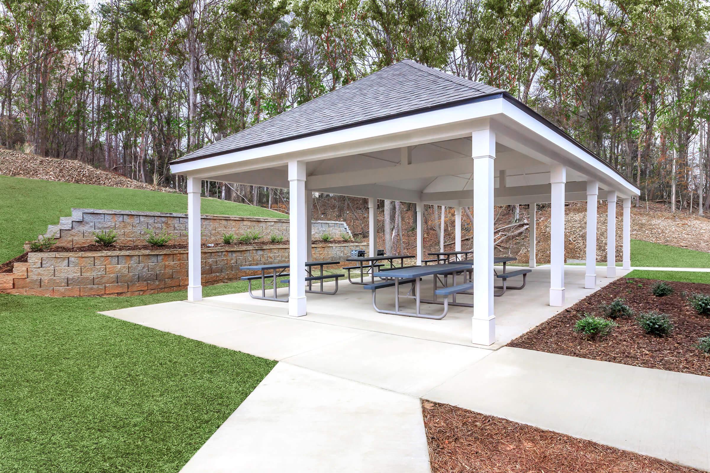 A covered outdoor pavilion with a sloped roof and white pillars, featuring multiple picnic tables underneath. Surrounding landscaping includes green grass, a stone retaining wall, and trees in the background. A paved walkway leads to the pavilion, creating a spacious and inviting area for gatherings.