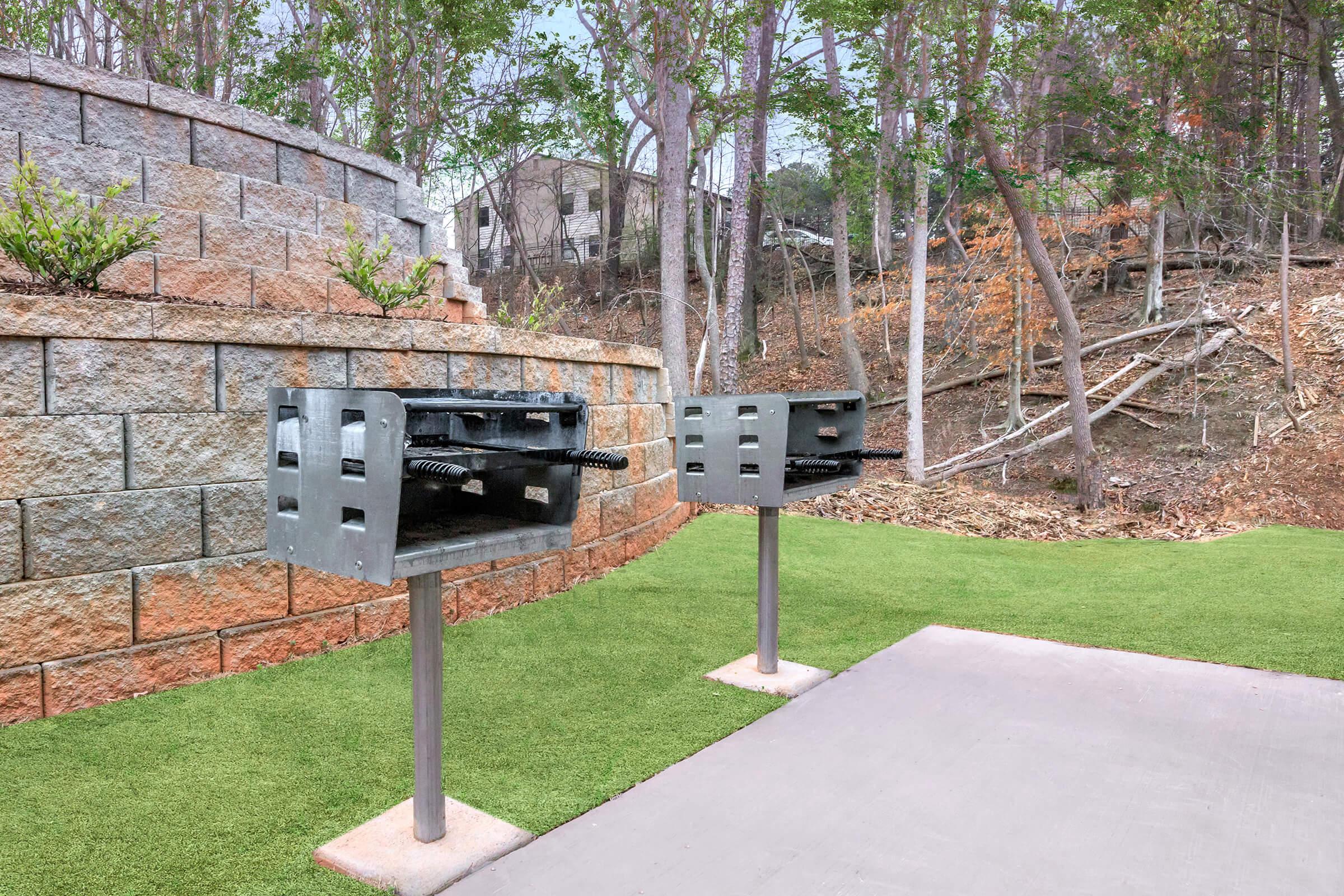 Two metal grilling stations with adjustable grates, mounted on vertical posts, positioned on a grassy area near a stone wall. Trees and a sloping landscape are visible in the background, providing a natural setting for outdoor cooking.