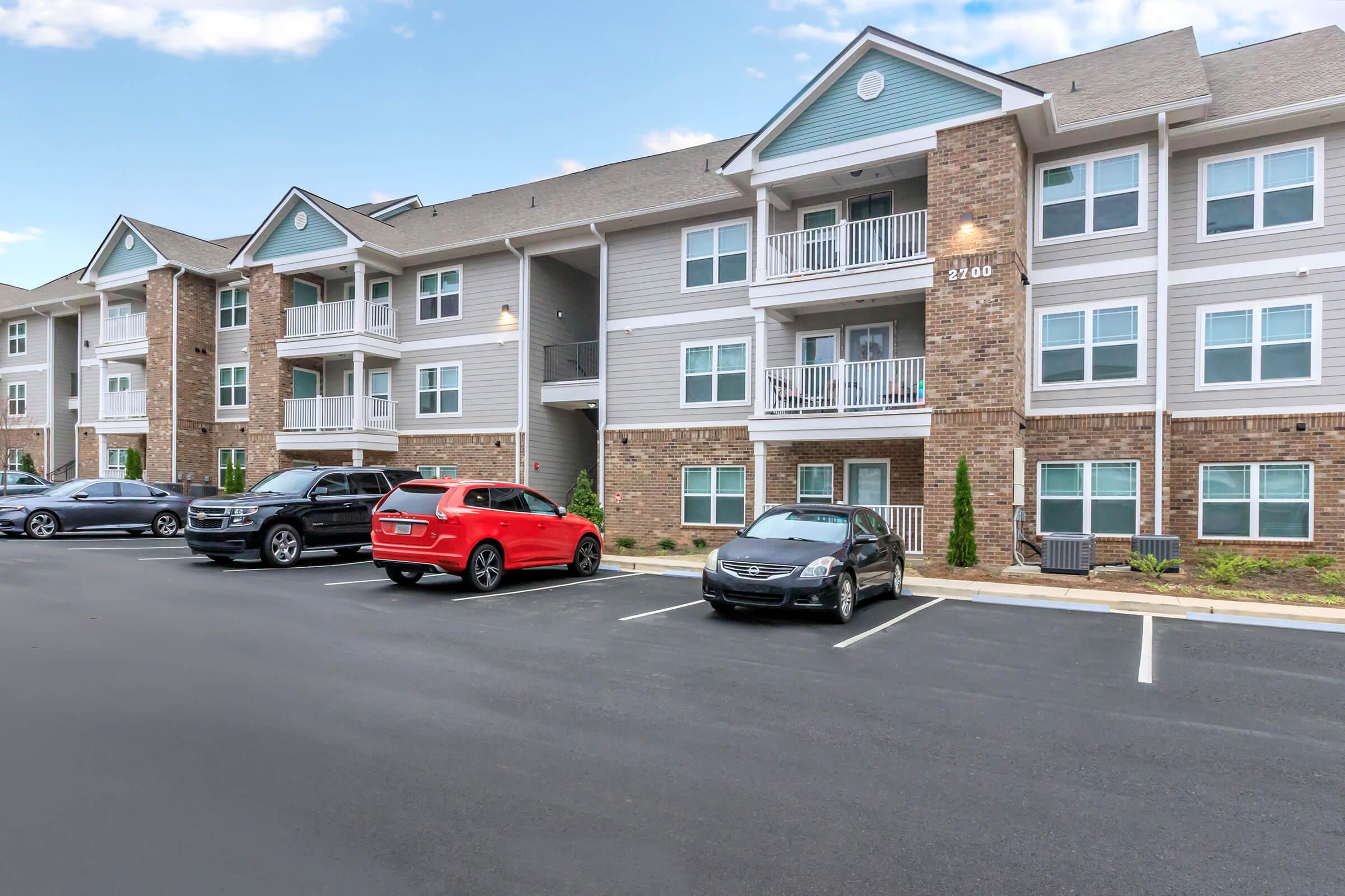 A modern apartment building with three stories, featuring brick and light-colored siding. Several parked cars, including a red SUV and a silver sedan, are visible in the foreground. The building has balconies and trees landscaping around it, set against a clear blue sky.