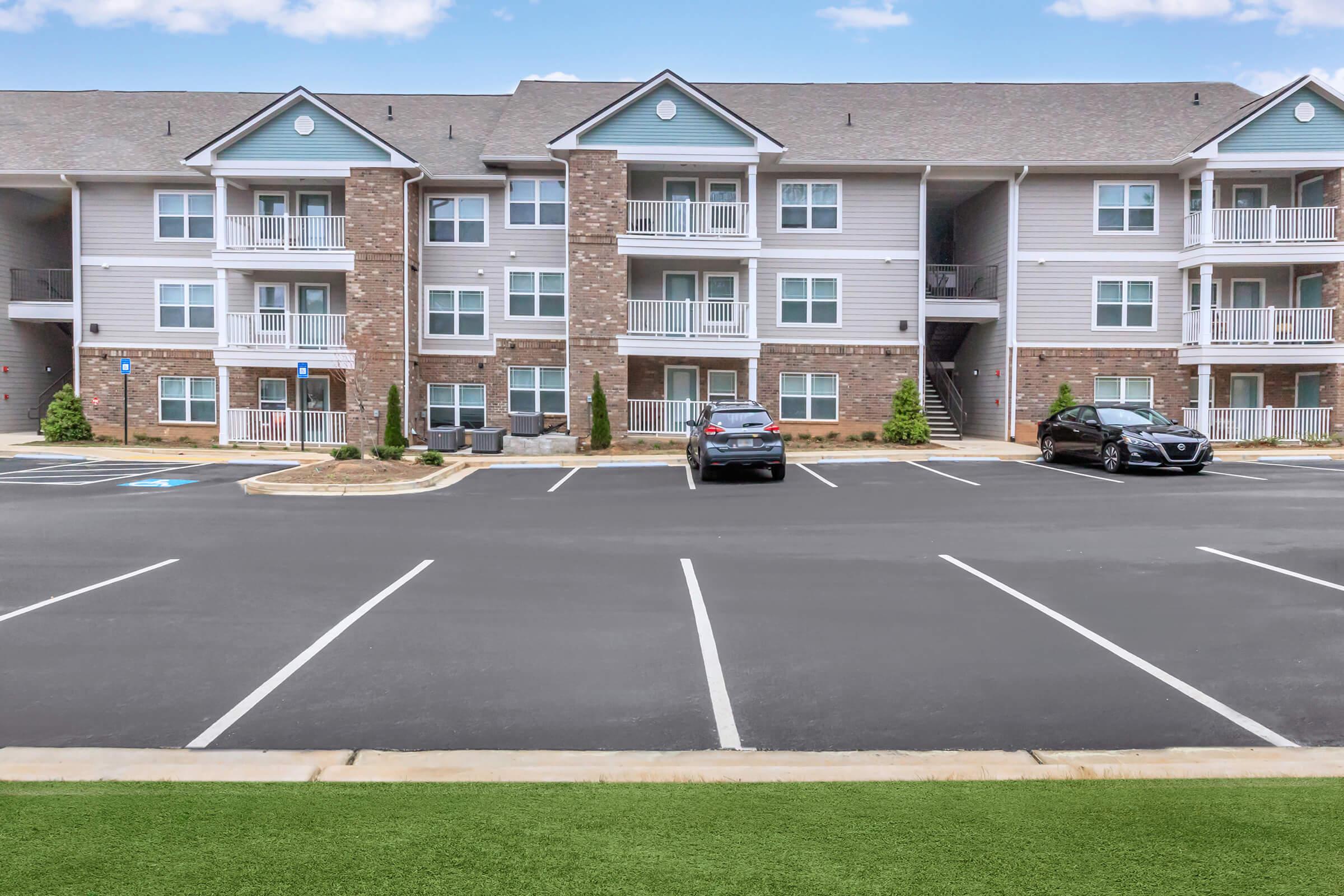 A multi-story apartment building featuring brick and siding exteriors. There are several parked cars in the foreground and a well-maintained green lawn. The building has balconies, and there are shrubs and small trees landscaping the entrance. The sky is clear with a few clouds.