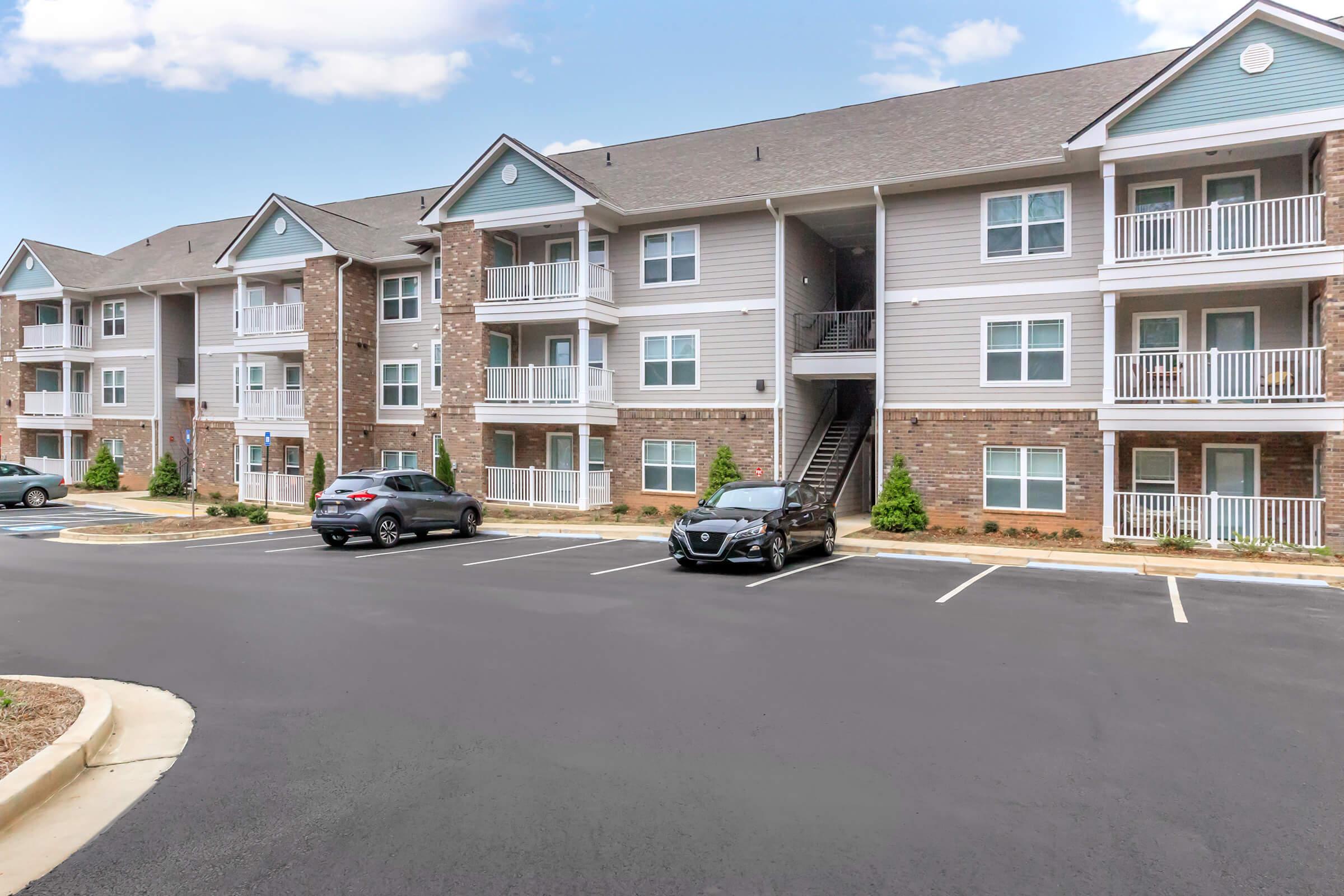 A multi-unit apartment building featuring three stories with brick and siding exteriors. The building has multiple balconies and adjacent parking spaces. There are several cars parked in front, and landscaping is visible around the building. The sky is partly cloudy.