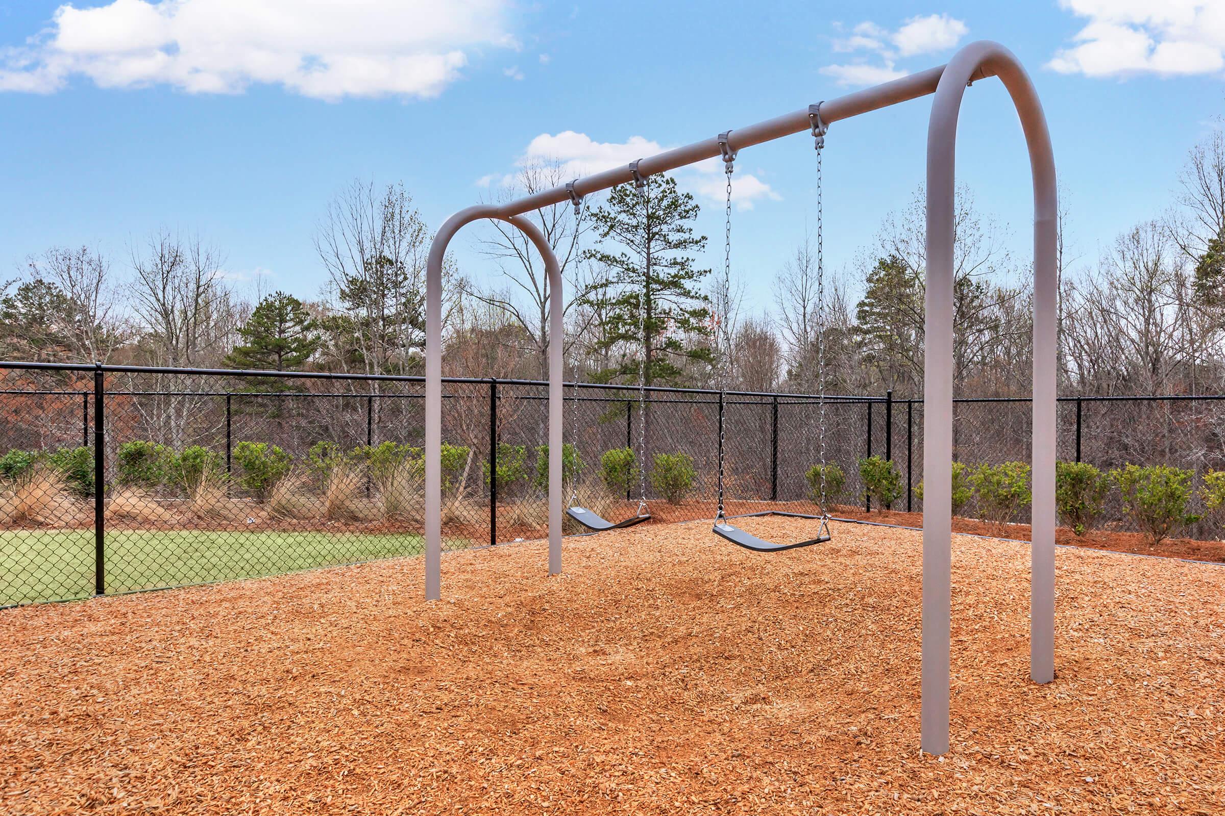 A playground scene featuring two empty swings on a metal frame, set on a bed of wood chips. The swings are surrounded by a chain-link fence, with trees in the background and a clear blue sky above. The area looks inviting and well-maintained, perfect for children to play.