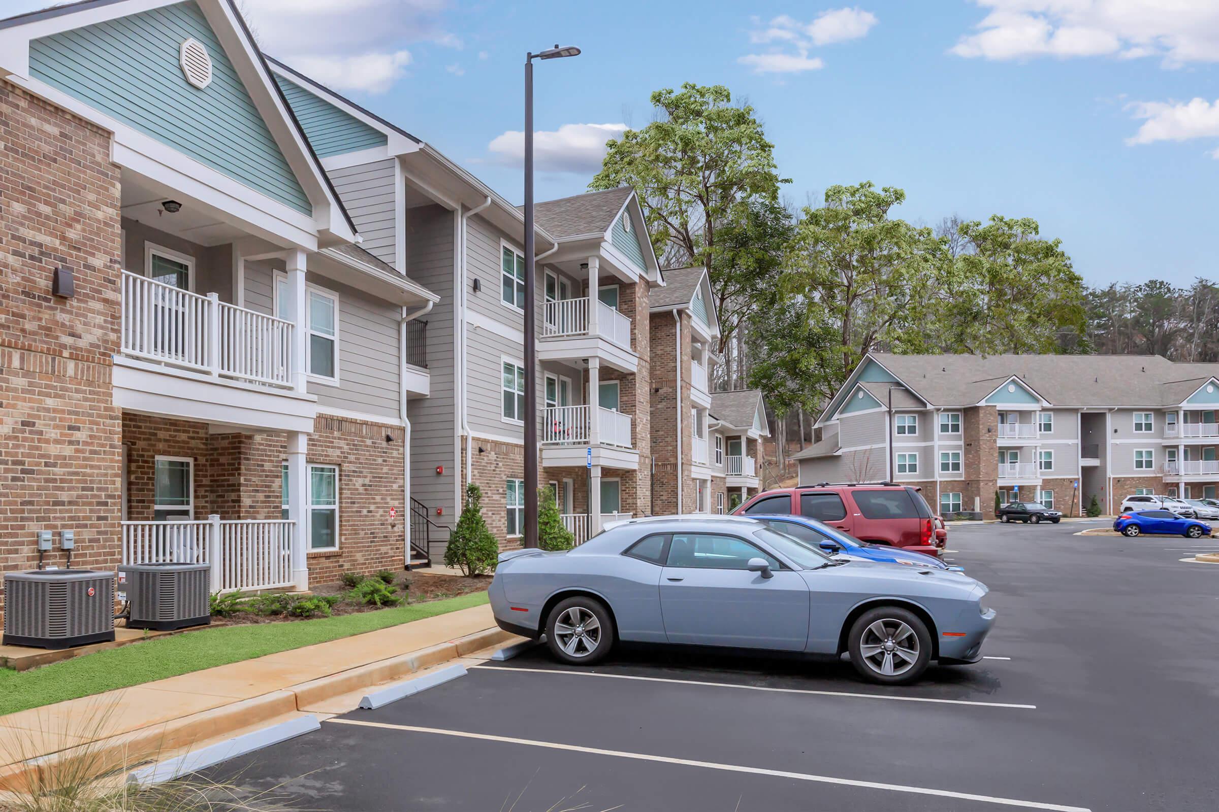 A residential apartment complex featuring two buildings with multiple units and balconies. In the foreground, a light blue car is parked next to a red SUV. The scene includes landscaped areas, parking spaces, and trees in the background under a partly cloudy sky.