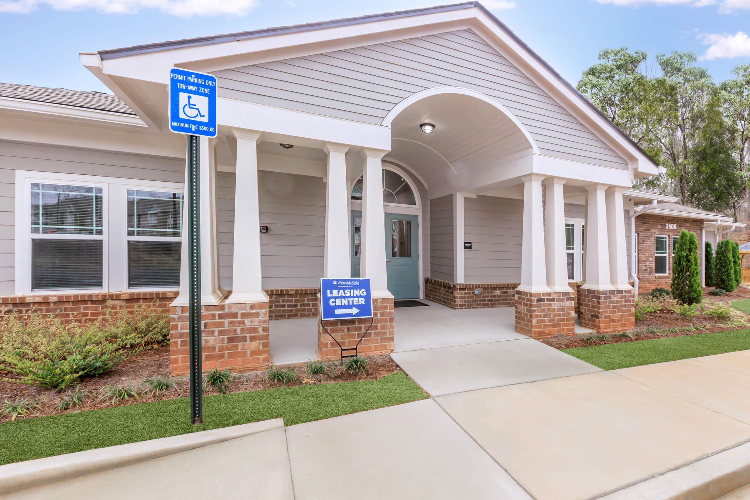 A modern leasing center with a welcoming entrance. The building features gray siding and brick accents, large windows, and a covered entryway. A blue sign indicates the leasing center location, and a parking sign designates accessible parking. Lush green landscaping surrounds the entrance, adding to the appealing atmosphere.