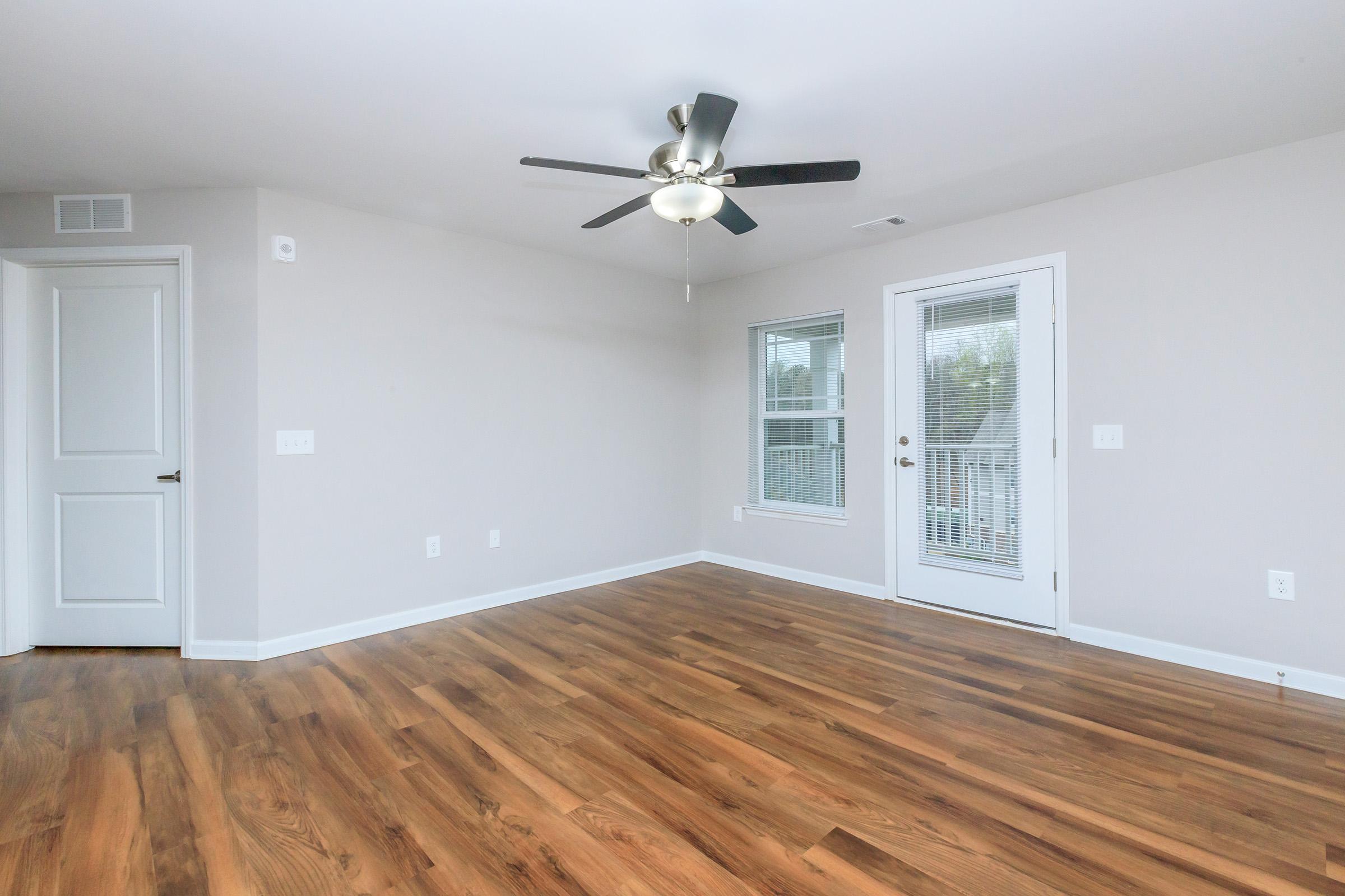 Spacious living room with light-colored walls and wood-style flooring. A ceiling fan is installed, and there's a sliding glass door leading to a balcony. Natural light fills the room, contributing to a bright and airy atmosphere. A small closet is visible near the entrance.