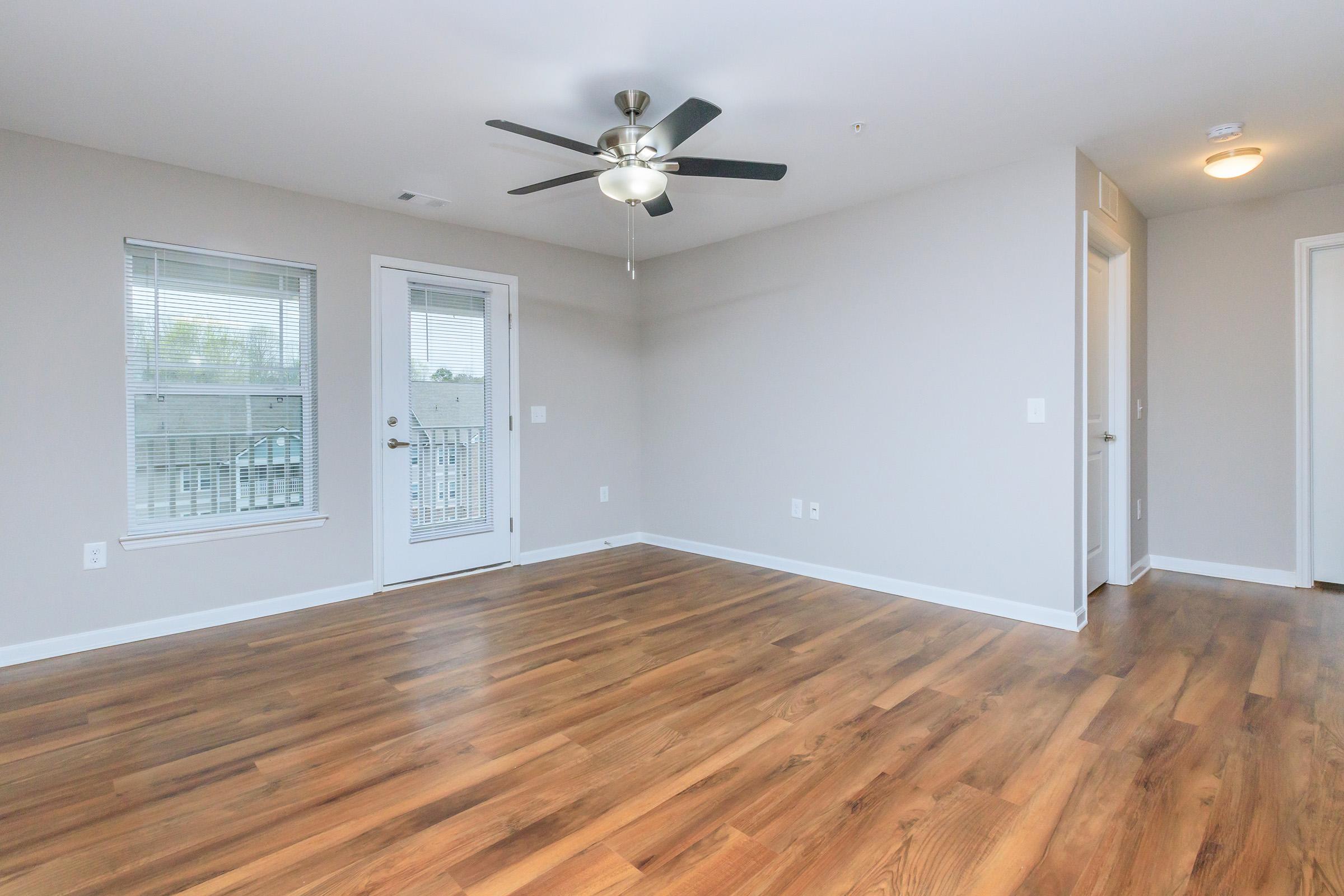 A spacious, empty room featuring neutral gray walls and a ceiling fan. The room has large windows allowing natural light, and a door leading to a balcony. The floor is made of light-colored hardwood, and there are white baseboards and door frames, creating a bright and airy atmosphere.