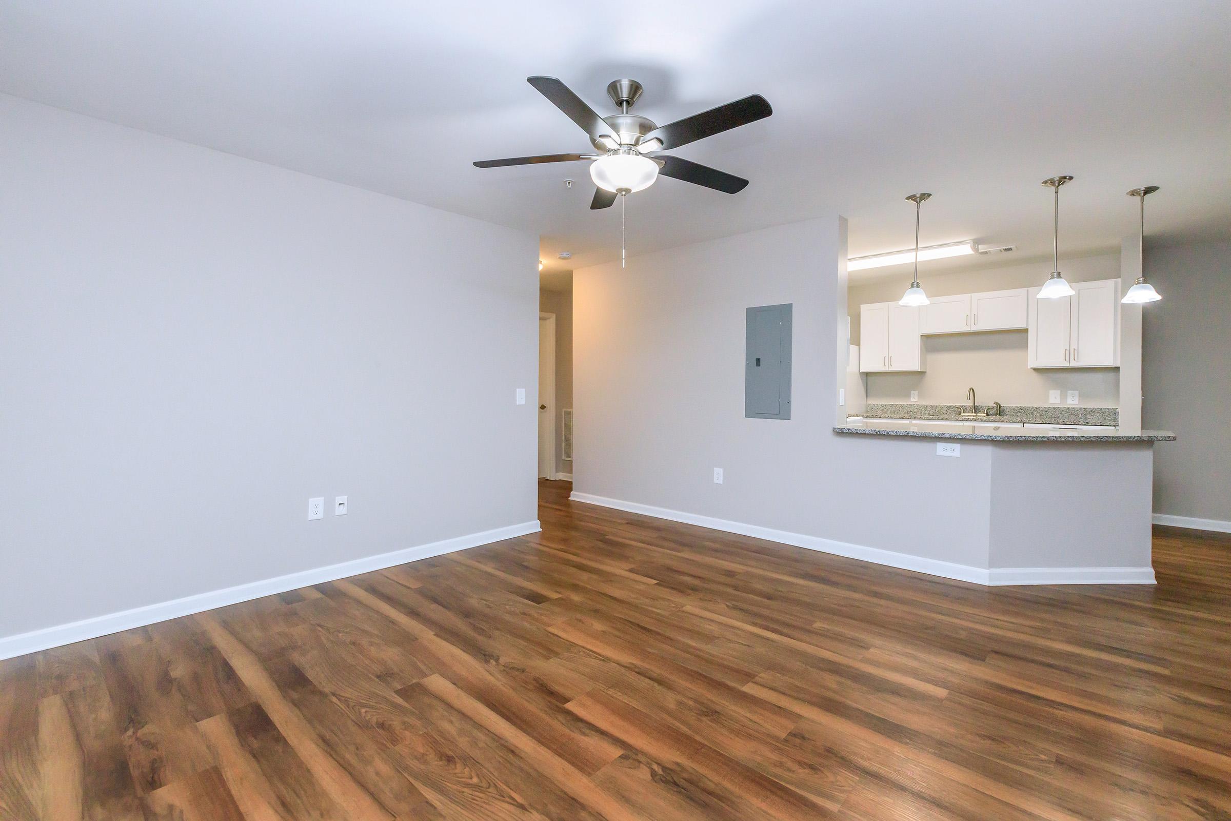 Spacious living area with light wood flooring, a ceiling fan, and neutral-colored walls. A kitchen with granite countertops and pendant lighting is visible in the background. The layout is open, creating a bright and inviting atmosphere.