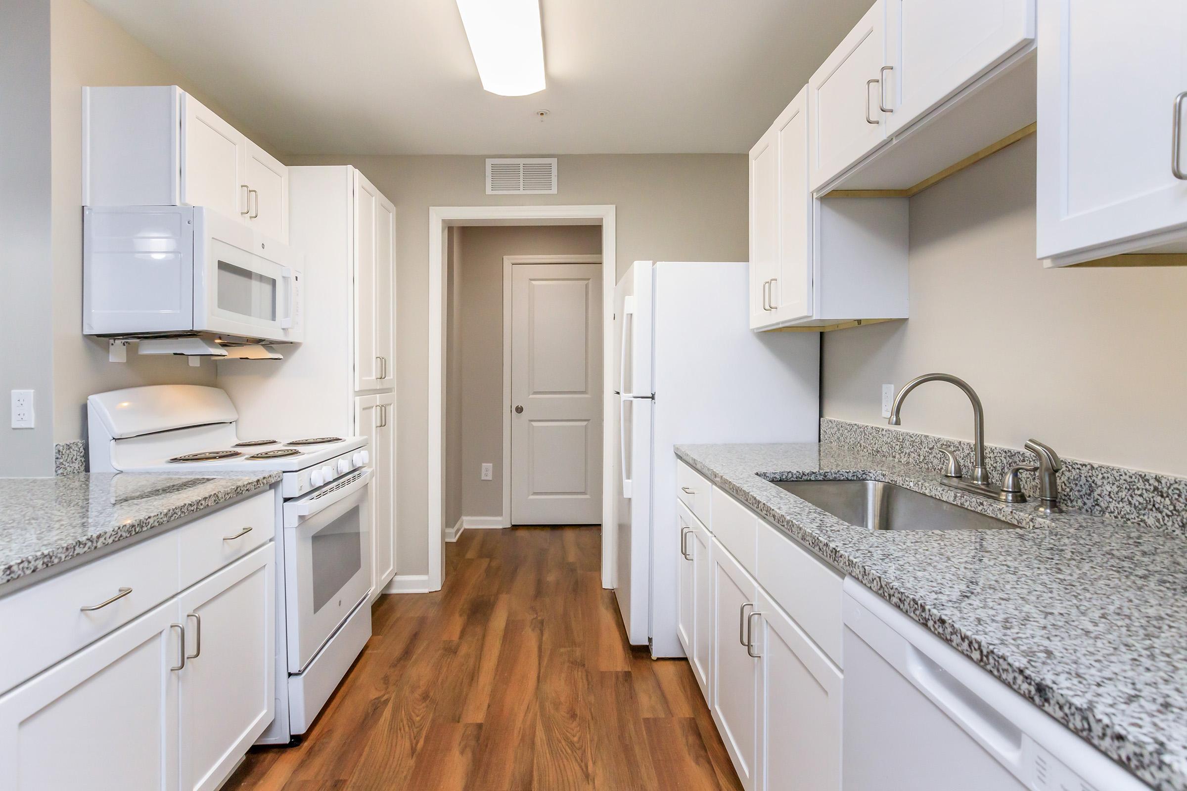Spacious modern kitchen featuring white cabinets, granite countertops, and stainless steel appliances. Includes a microwave, oven, and refrigerator. Well-lit with overhead lighting, and warm wood flooring. A doorway leads to another room.