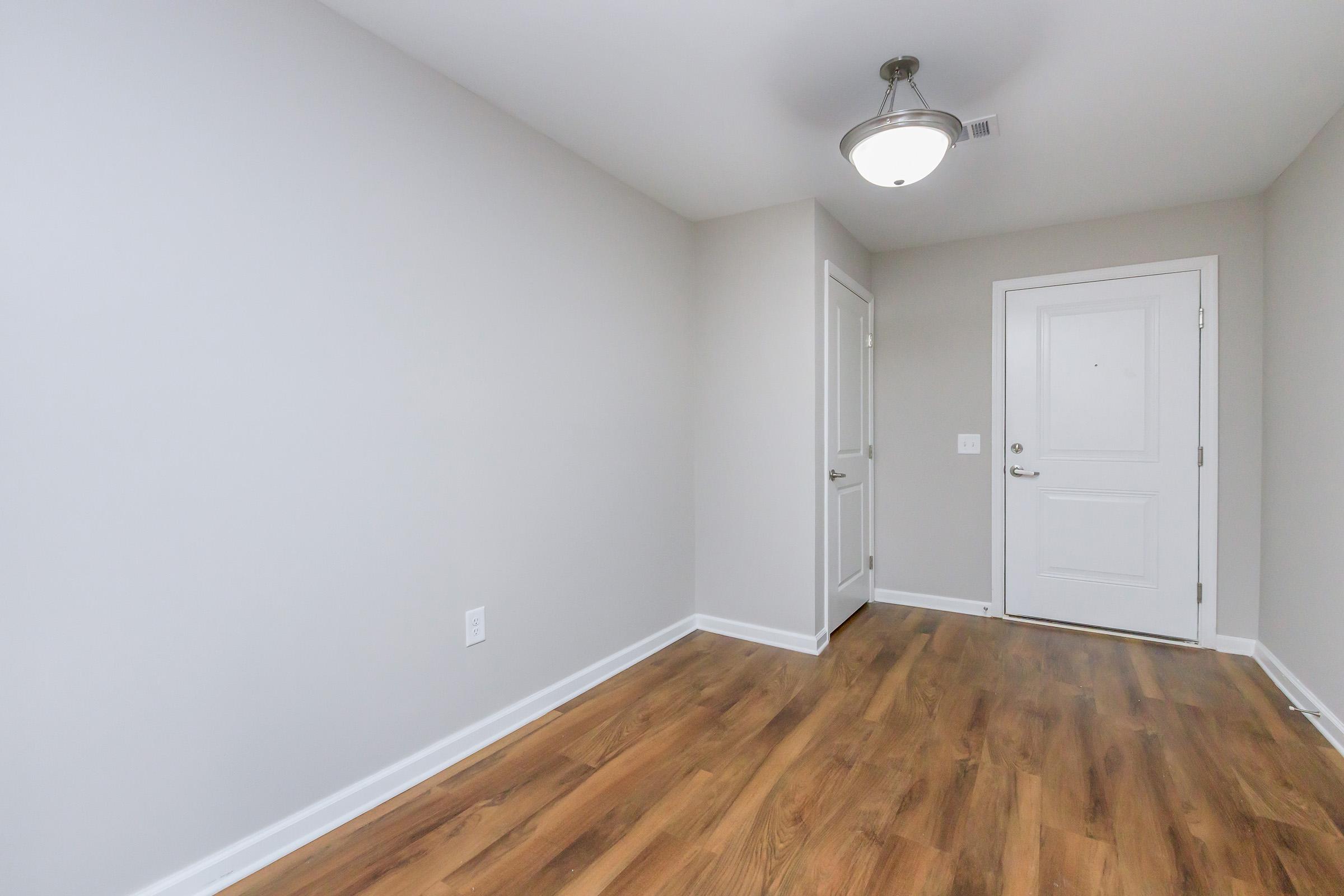 A well-lit, empty entrance hallway with light gray walls and a wooden floor. The space features a white door with a doorknob, a closet door, and a ceiling light fixture. The overall aesthetic is clean and modern, creating an inviting atmosphere.