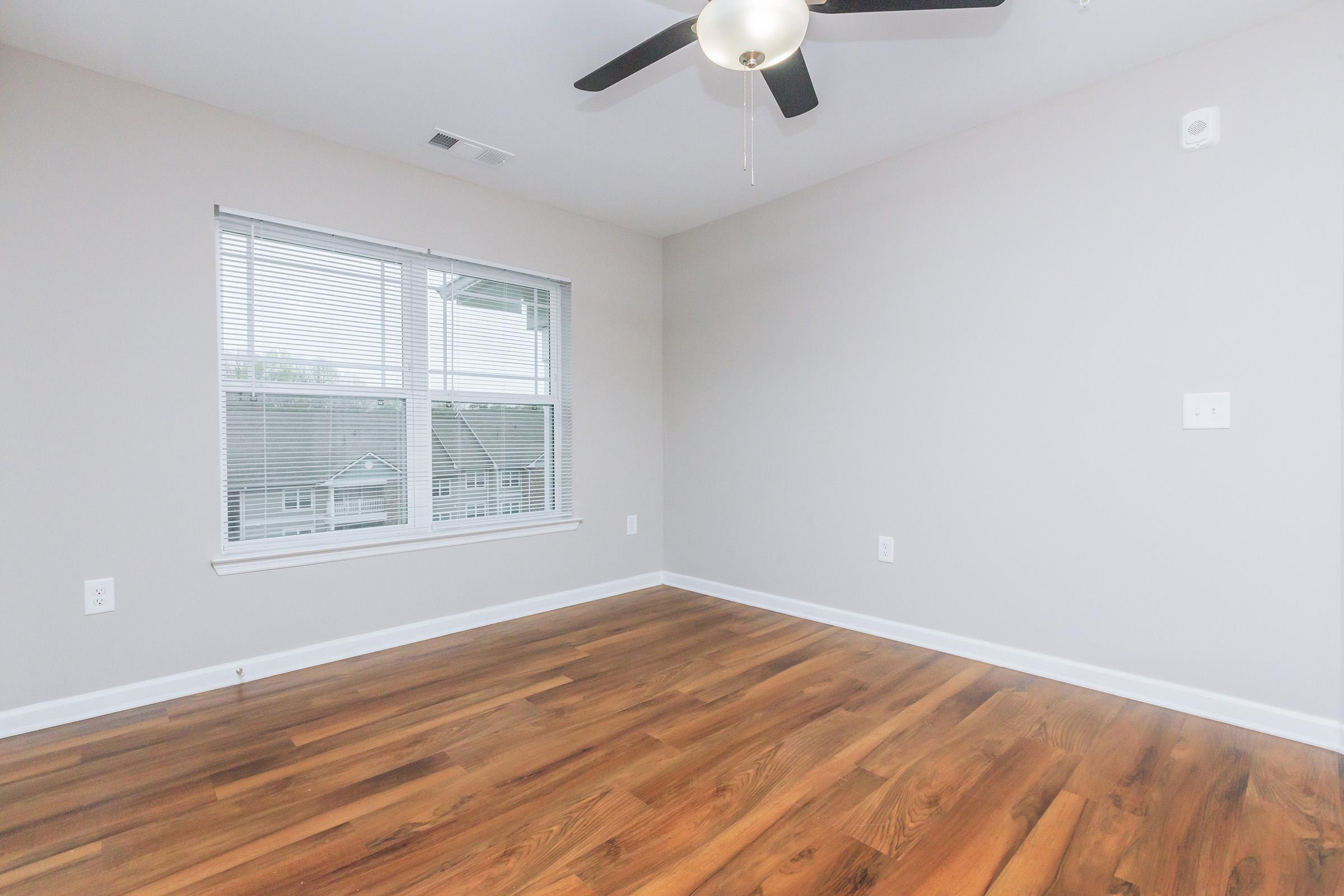 A clean, empty room with light gray walls and brown wooden flooring. There is a ceiling fan with three blades, and a window with blinds allowing natural light in. The room appears spacious and is freshly painted, providing a blank canvas for furnishing.