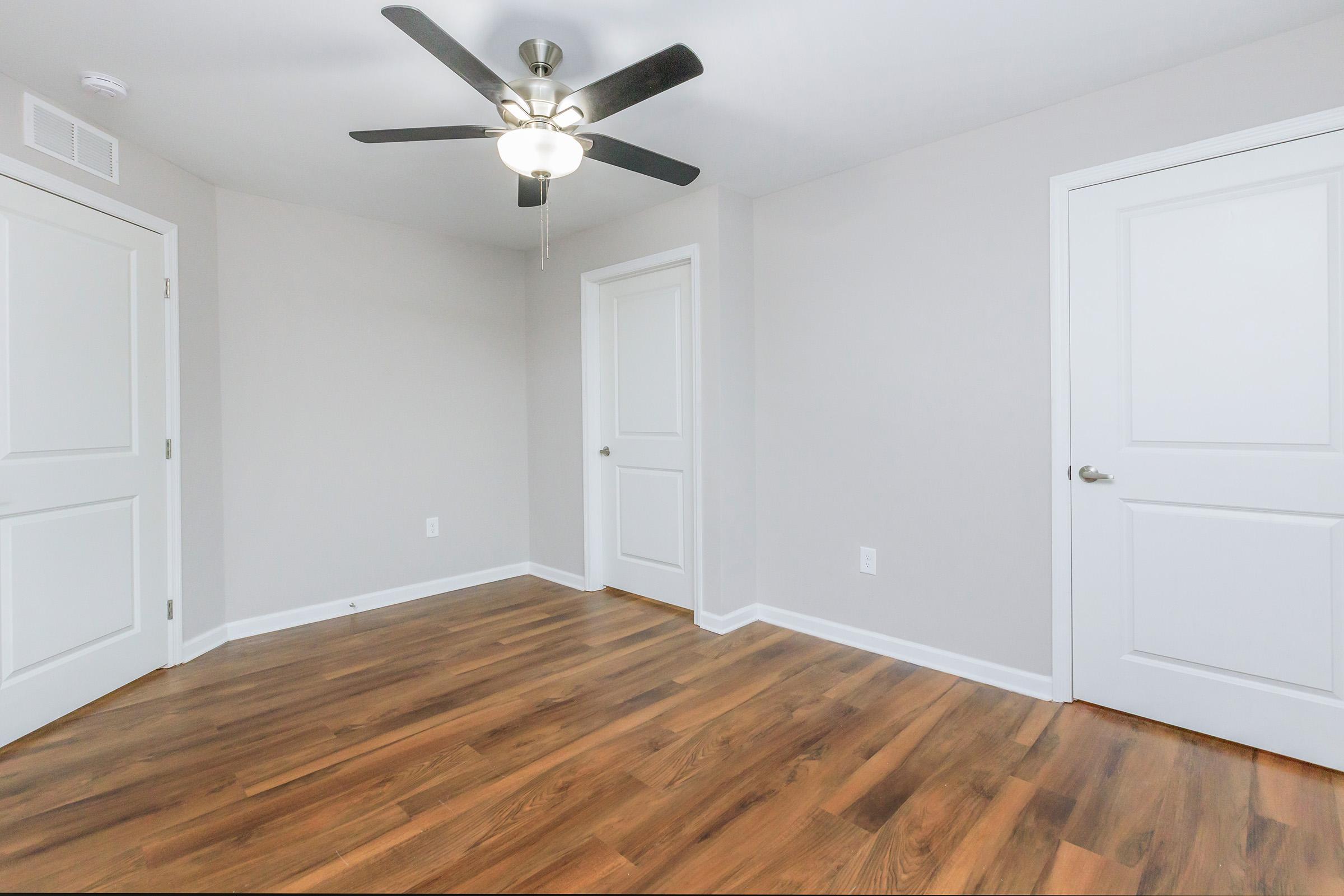 A well-lit empty room featuring light gray walls, a ceiling fan, and two closed white doors. The floor is covered in dark wood laminate, providing a warm contrast to the walls. The room has a clean and modern appearance, with no furniture or decorations present.