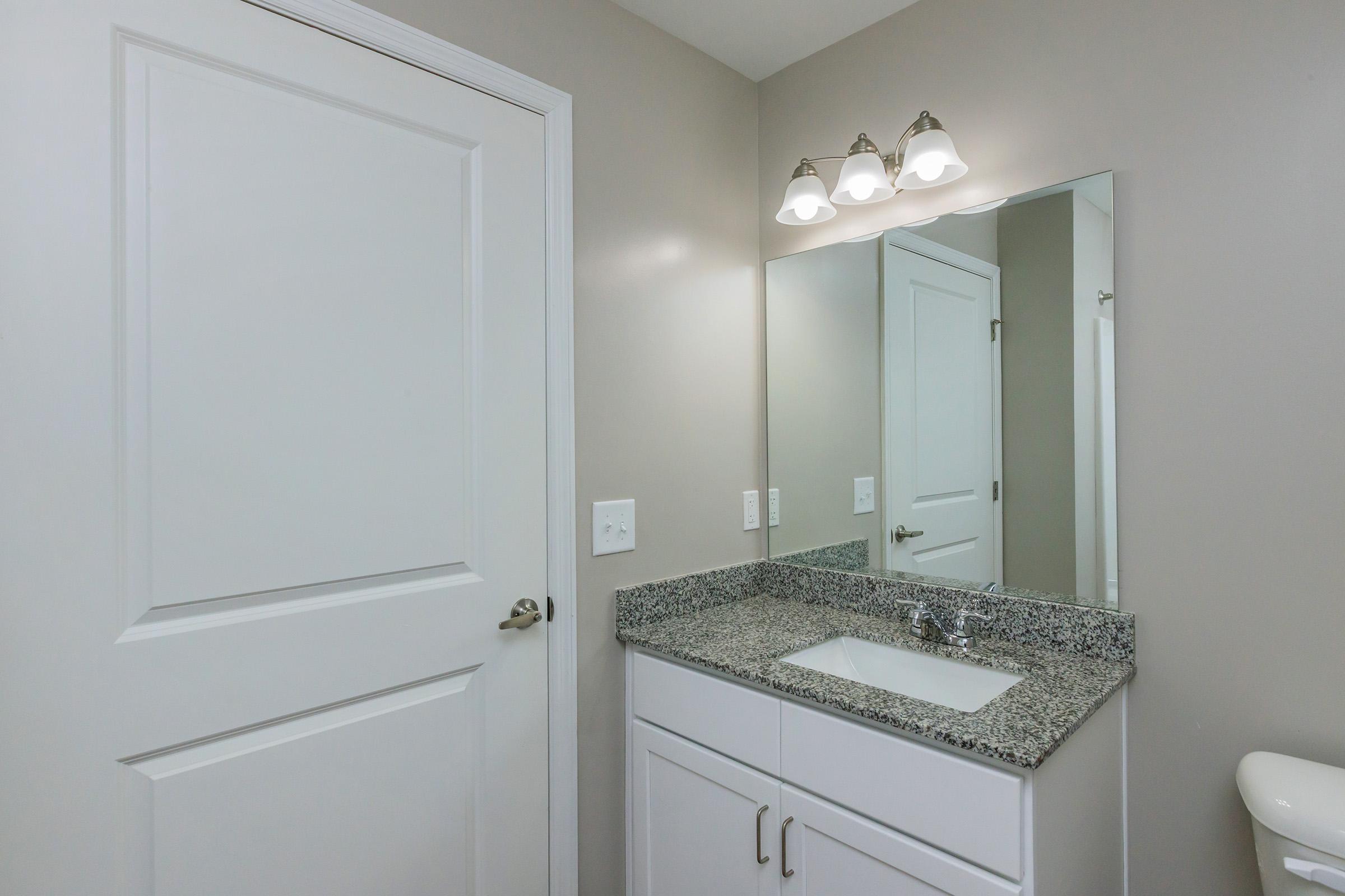 A modern bathroom featuring a white vanity with a granite countertop, a mirrored wall above the sink, and three light fixtures. The wall is painted in a neutral tone, and there's a white door partially visible on the left side of the image. The overall design is clean and contemporary.