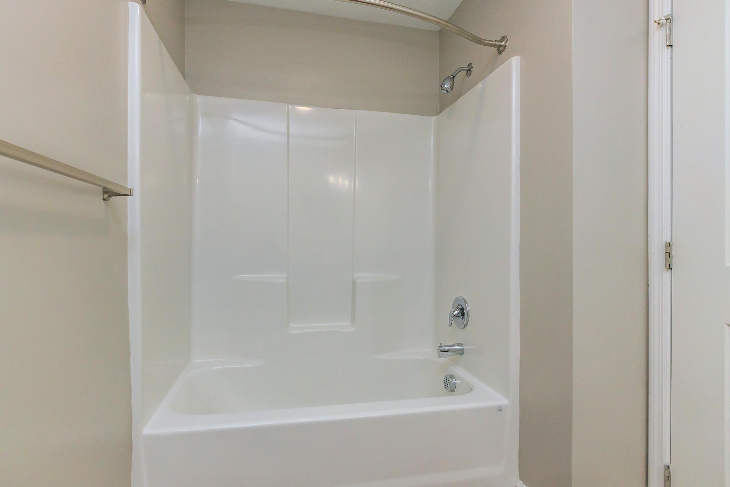 A clean, empty bathroom shower with a white tub surround, a chrome faucet, and a handheld showerhead. The walls are painted a light beige, and there is a towel bar installed on the side. The image focuses on the interior of the shower space.