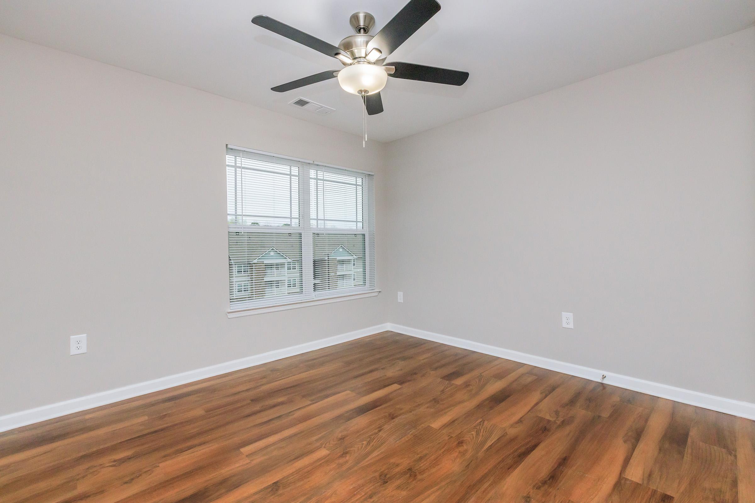 Empty room with light-colored walls and hardwood flooring. A ceiling fan with five blades is mounted in the center. There is a window with multiple panes allowing natural light to enter, and a scenic view outside. The space is uncluttered and ready for furnishing.