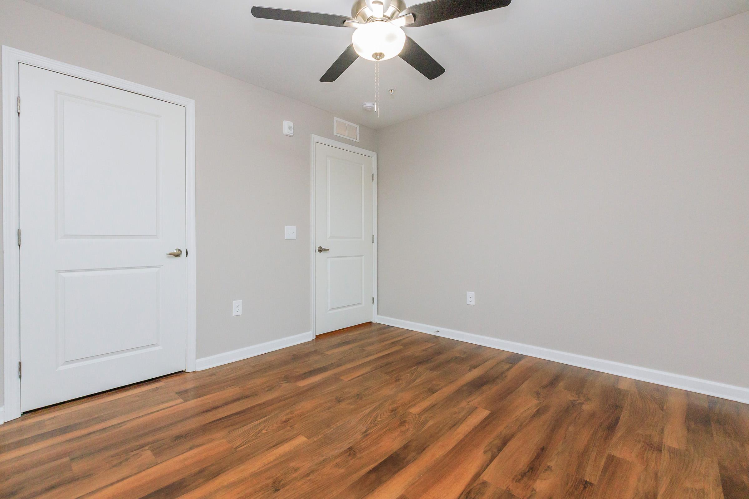 Empty room with light-colored walls and wooden flooring. A ceiling fan is visible, and there are two doors—one slightly ajar. The space is well-lit and uncluttered, creating a clean, minimalistic atmosphere.